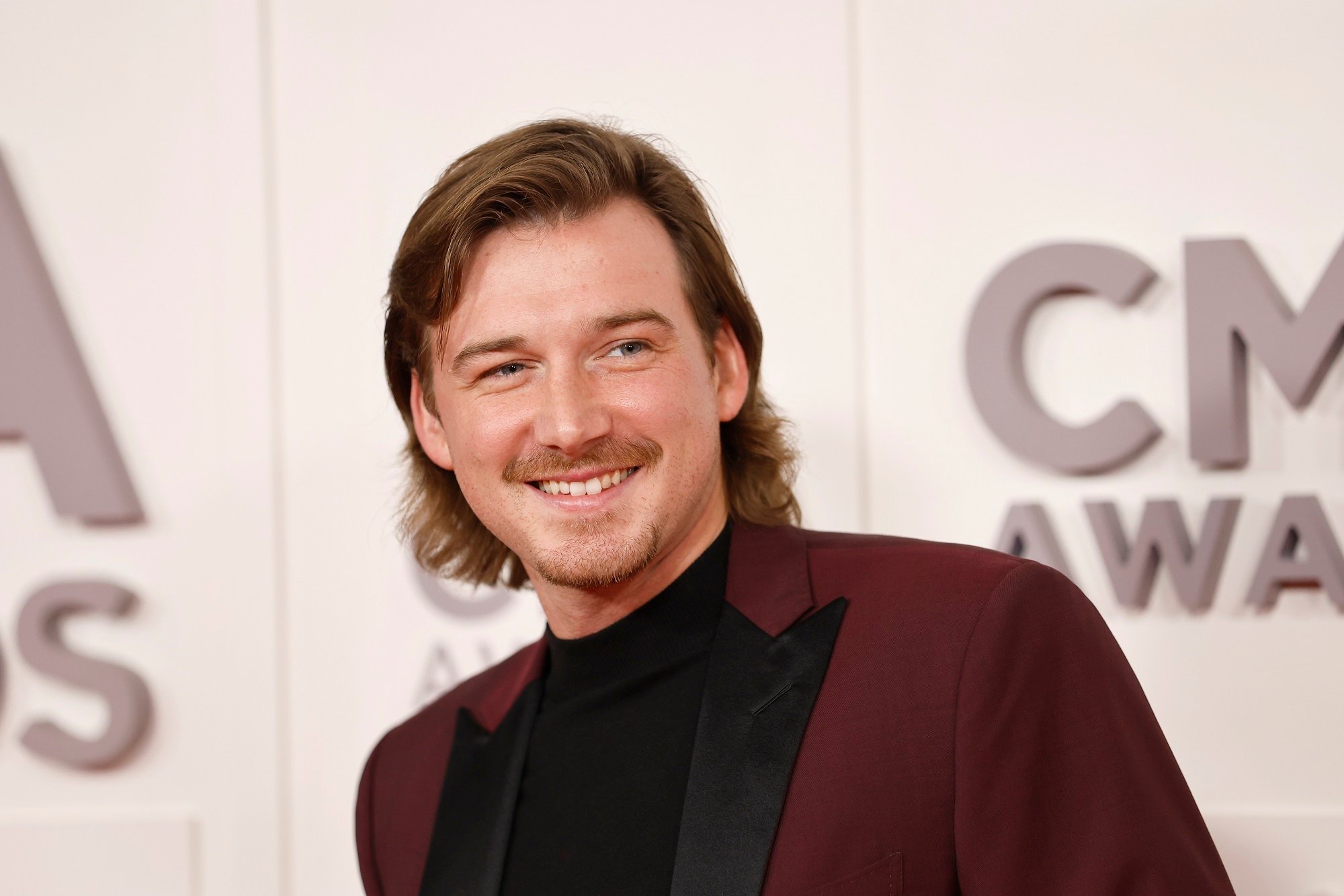 Morgan Wallen smiles while wearing a burgundy and black suit