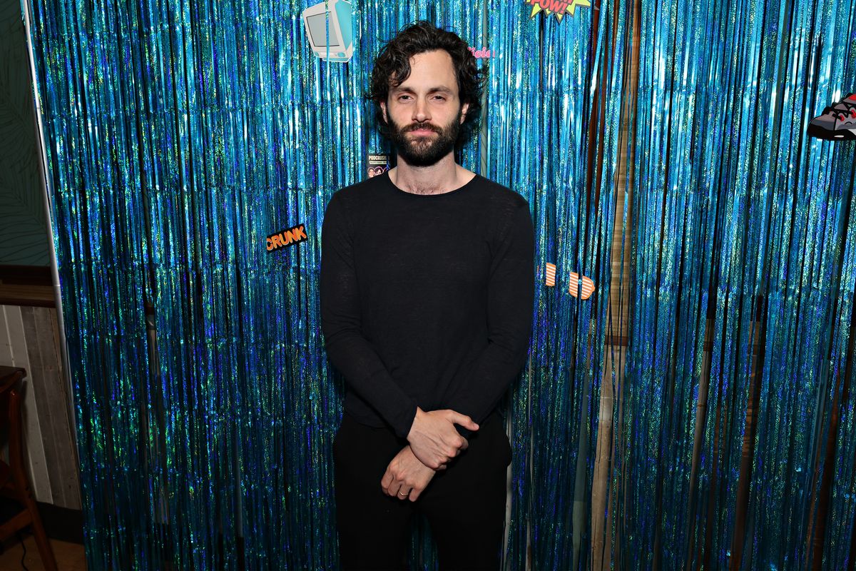 Penn Badgley poses in front of a shimmering blue backdrop made with streamers.