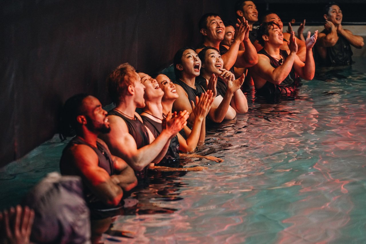 The 'Physical 100' cast stand in a pool and cheer looking up.