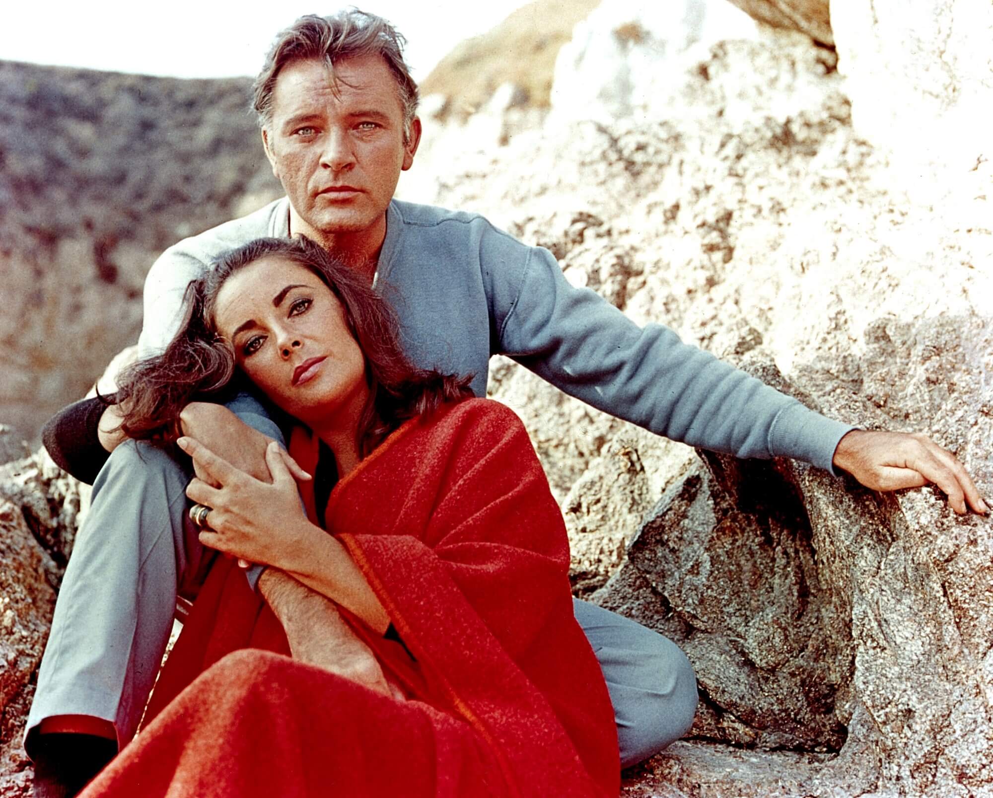 Richard Burton and Elizabeth Taylor sit outside on rocky ground