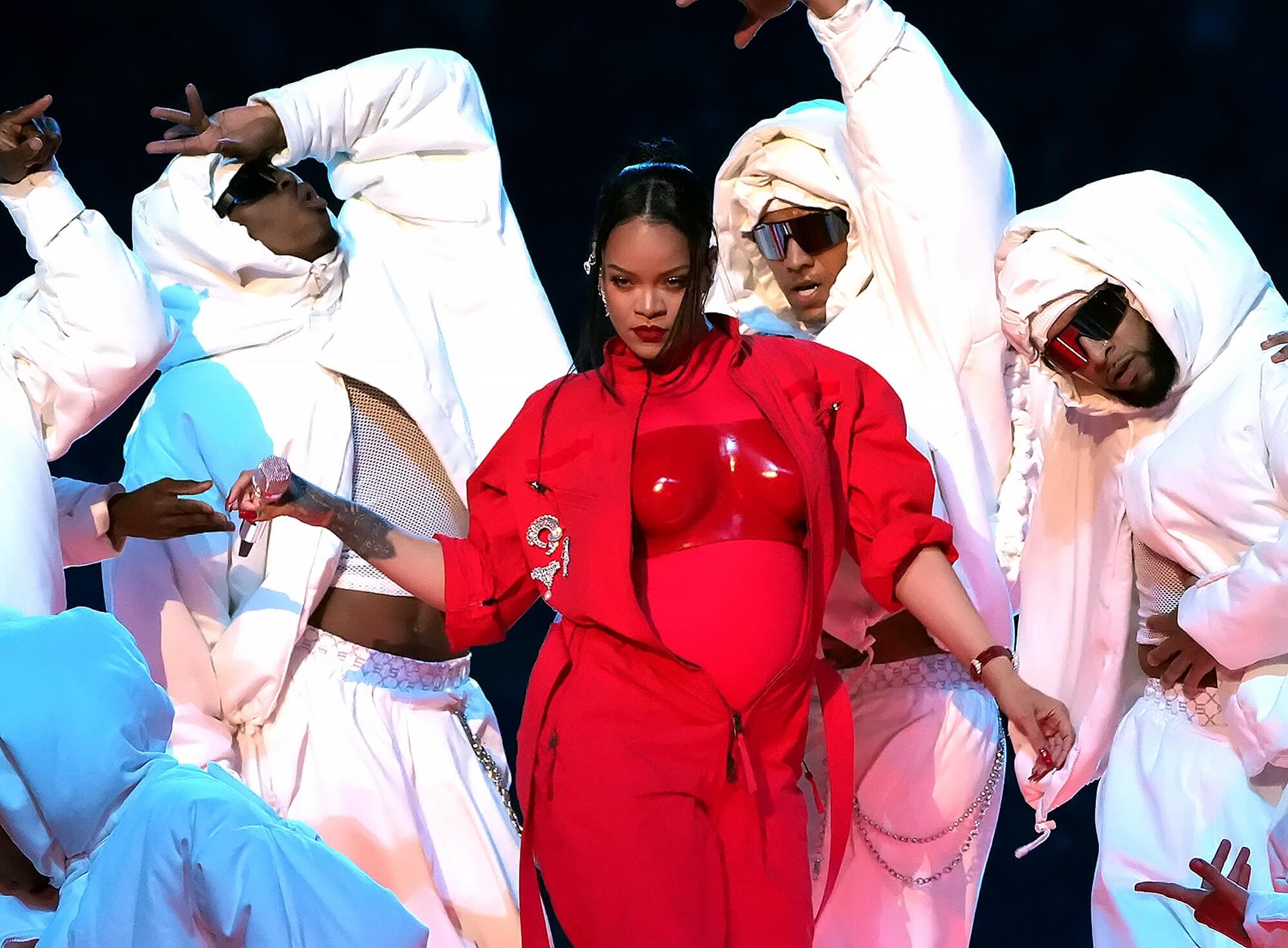 Rihanna wears a red outfit while surrounded by backup dancers in white outfits