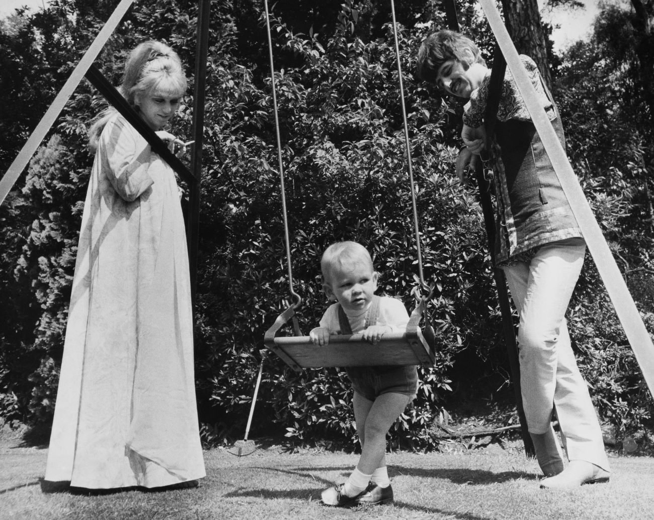 A black and white picture of Maureen Starkey and Ringo Starr looking at their son Zak on a swing.