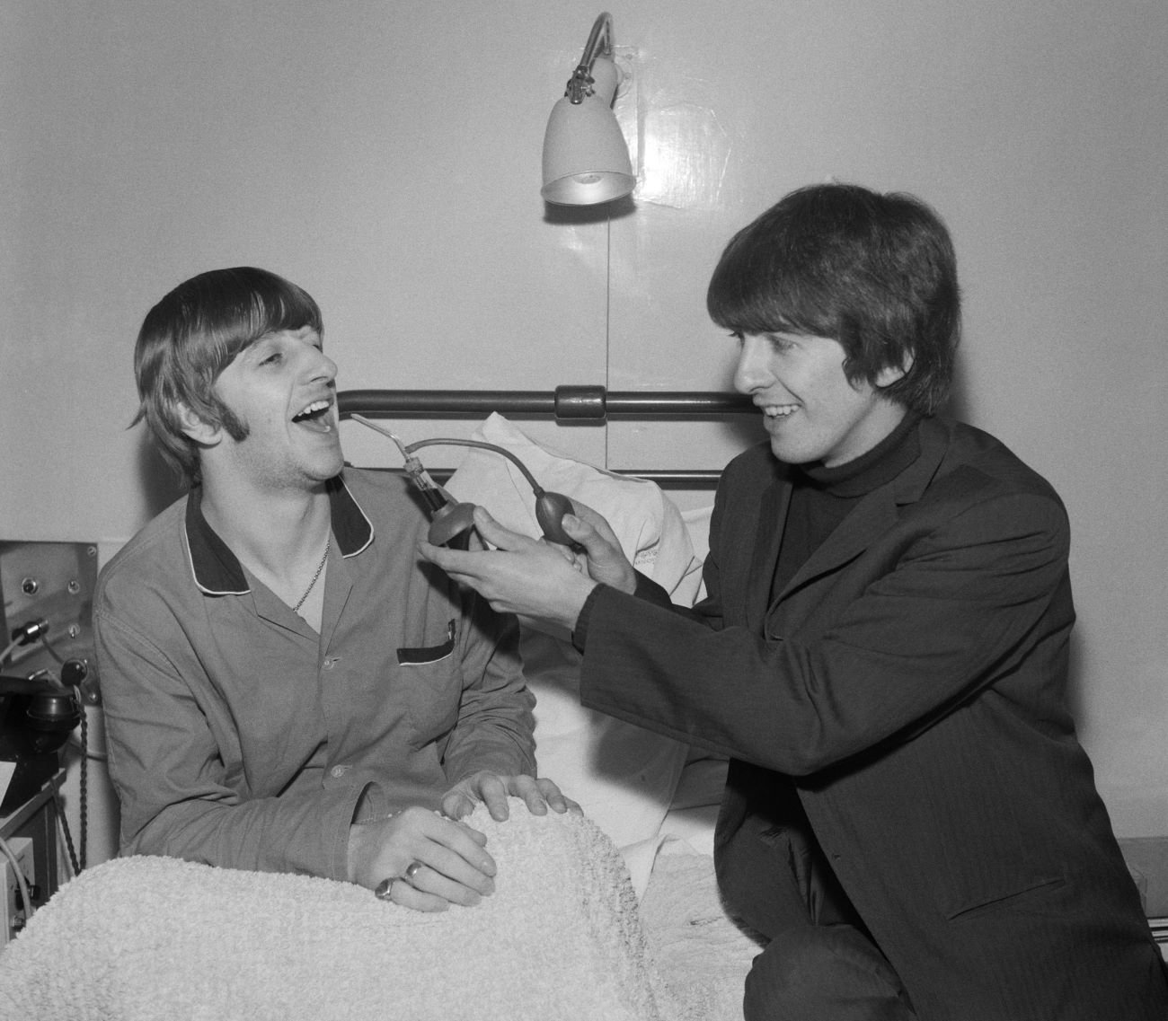 A black and white picture of Ringo Starr sitting in a bed and George Harrison sitting at his bedside. 