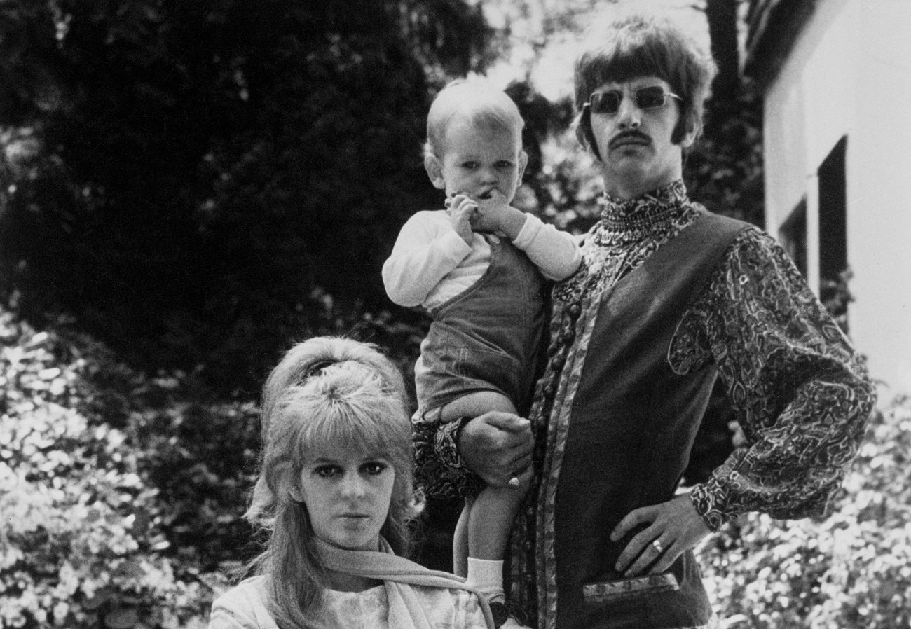 A black and white picture of Maureen Starkey sitting and Ringo Starr standing and holding his son, Zak Starkey.