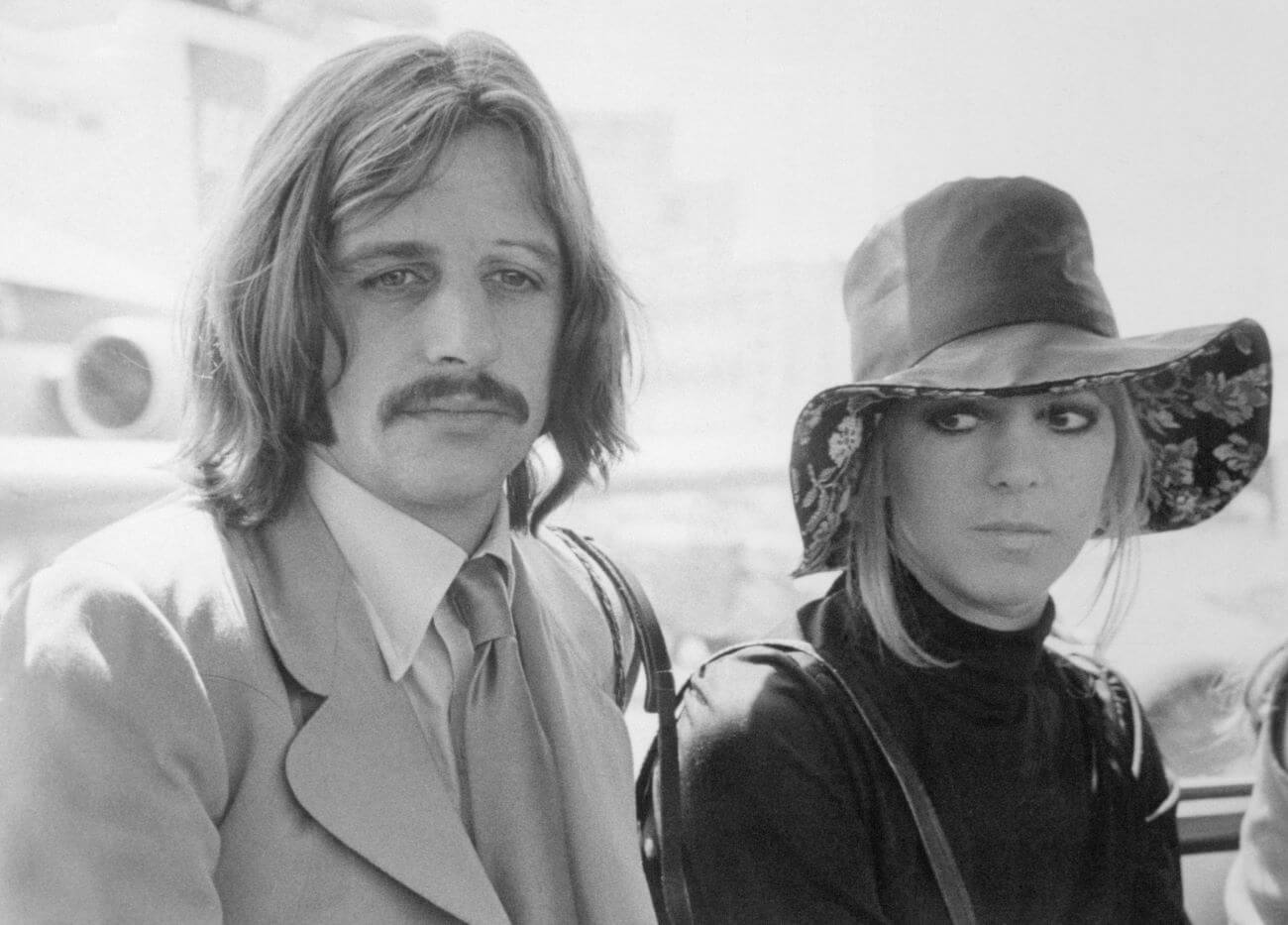 A black and white picture of Ringo Starr sitting next to his wife, Maureen Starkey, who wears a hat.