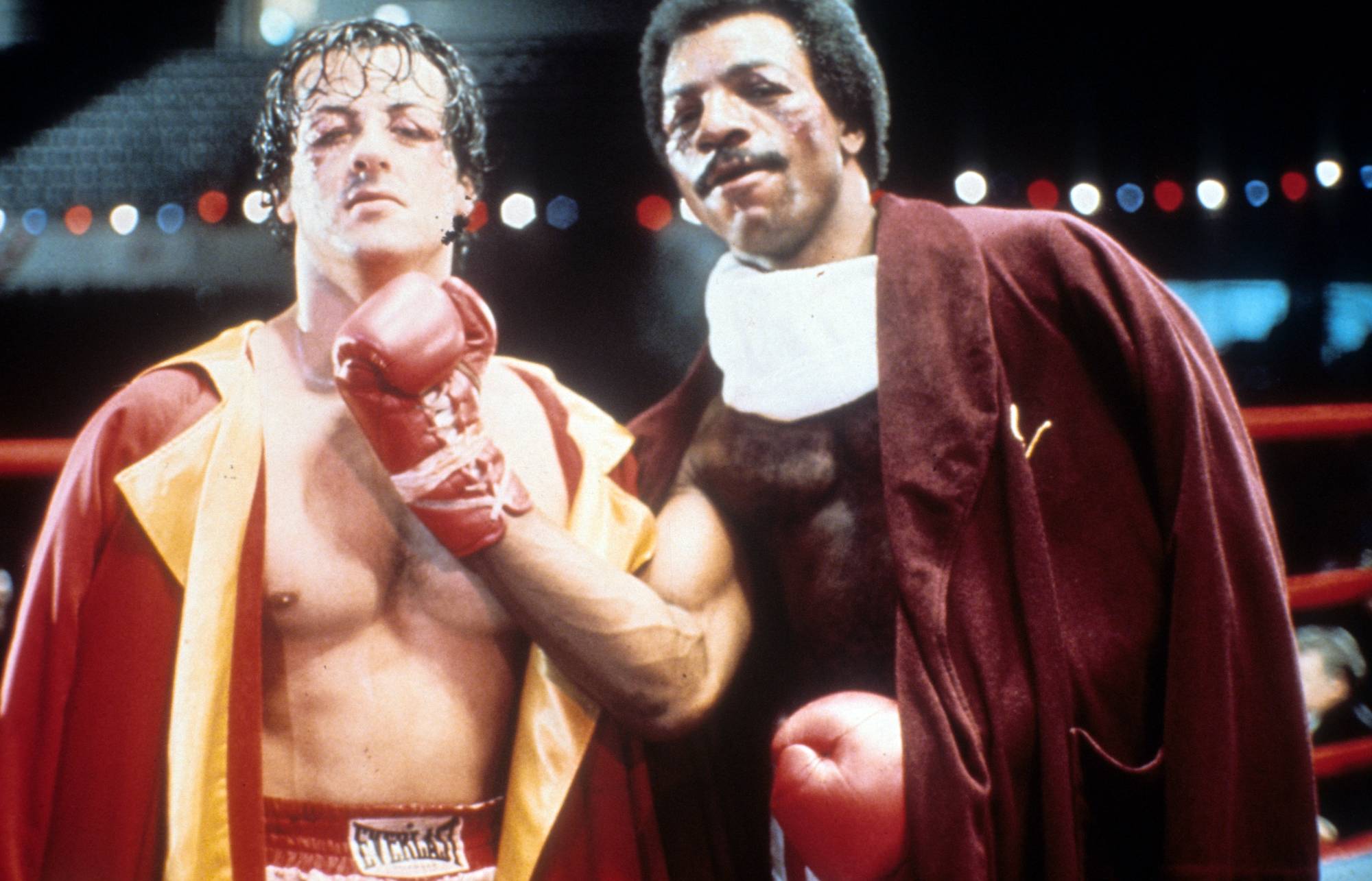 'Rocky' Sylvester Stallone as Rocky and Carl Weathers as Apollo Creed. Apollos is holding his boxing glove under Rocky's chin while they're wearing robes.