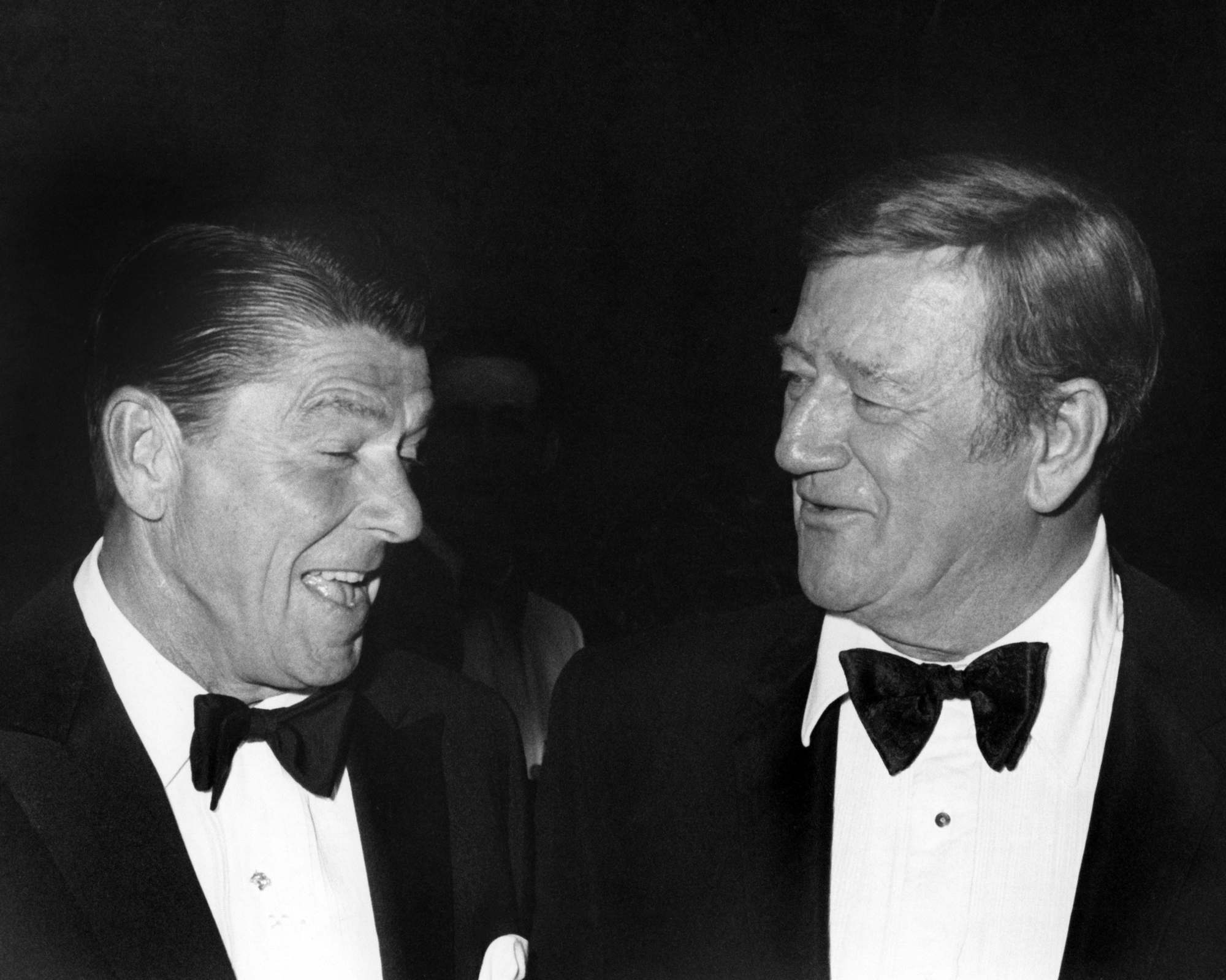 Ronald Reagan and John Wayne wearing tuxedos and talking in a black-and-white picture