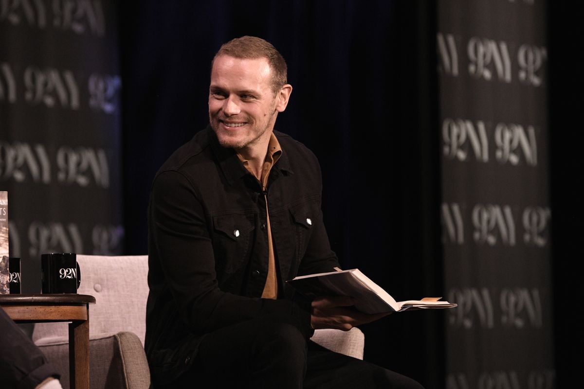 Sam Heughan smiles while holding a book.
