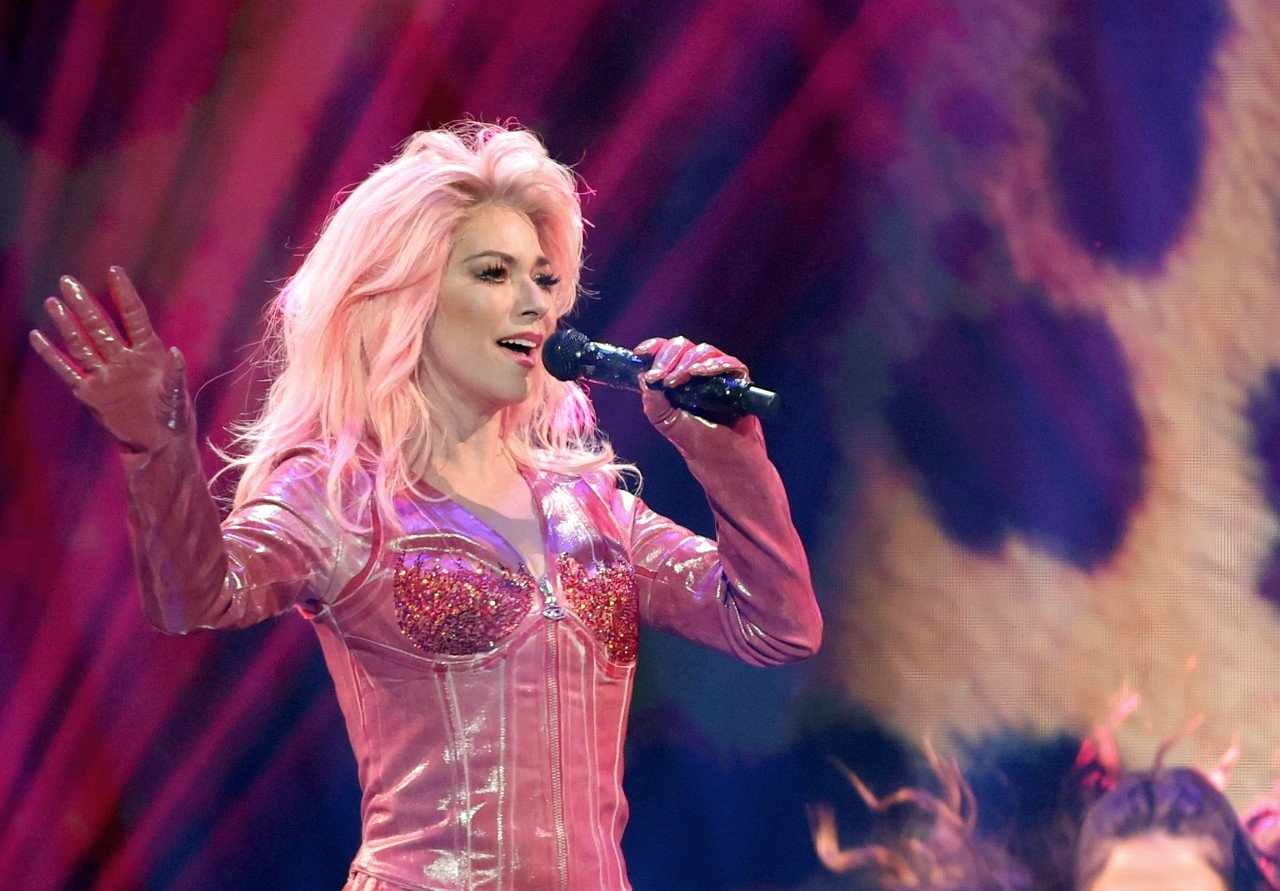Shania Twain poses in a pink outfit while singing at a concert.