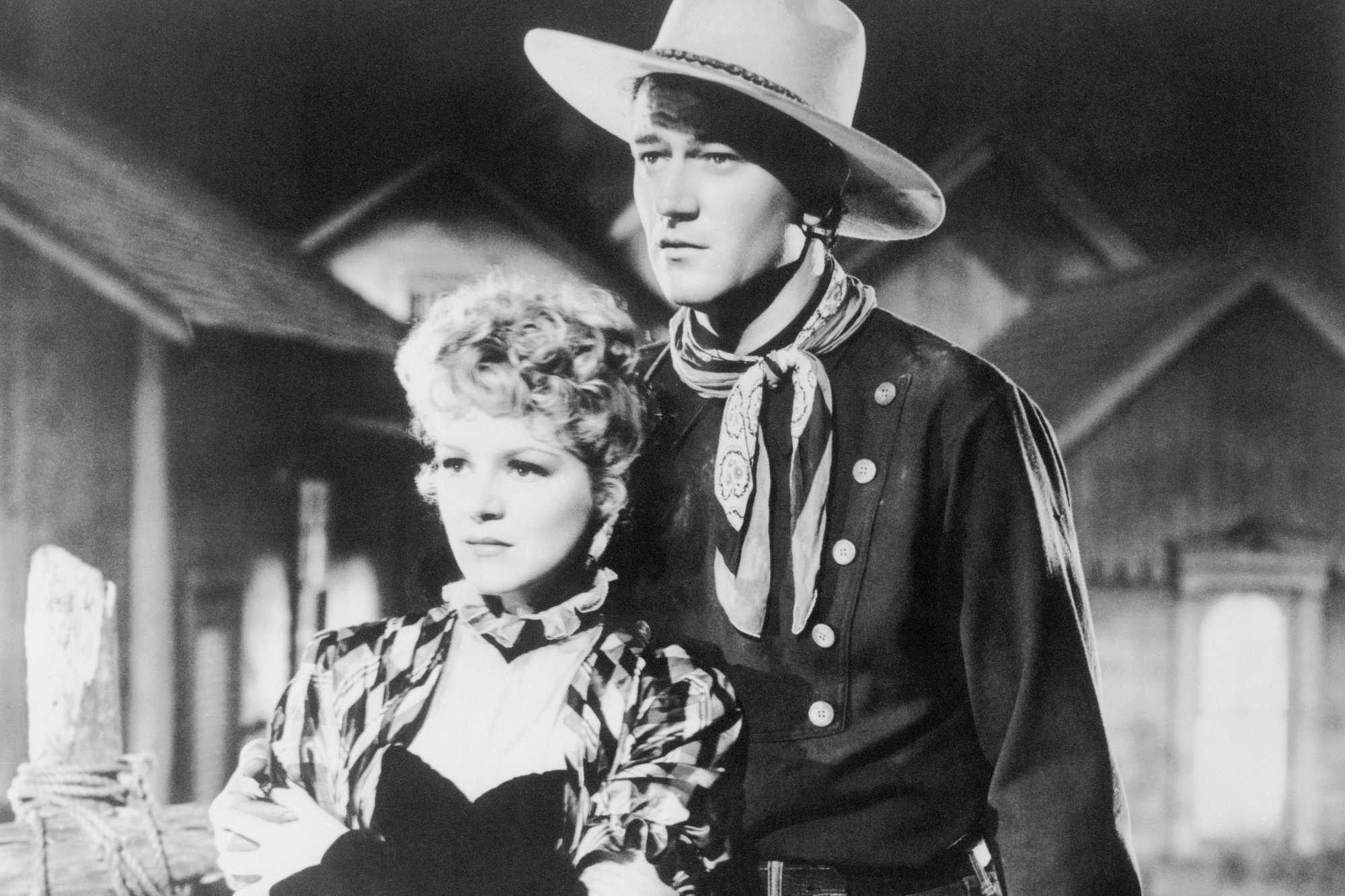 'Stagecoach' Claire Trevor as Dallas and John Wayne as Ringo Kid in John Ford movie. They're standing next to one another in Western costumes looking surprised.