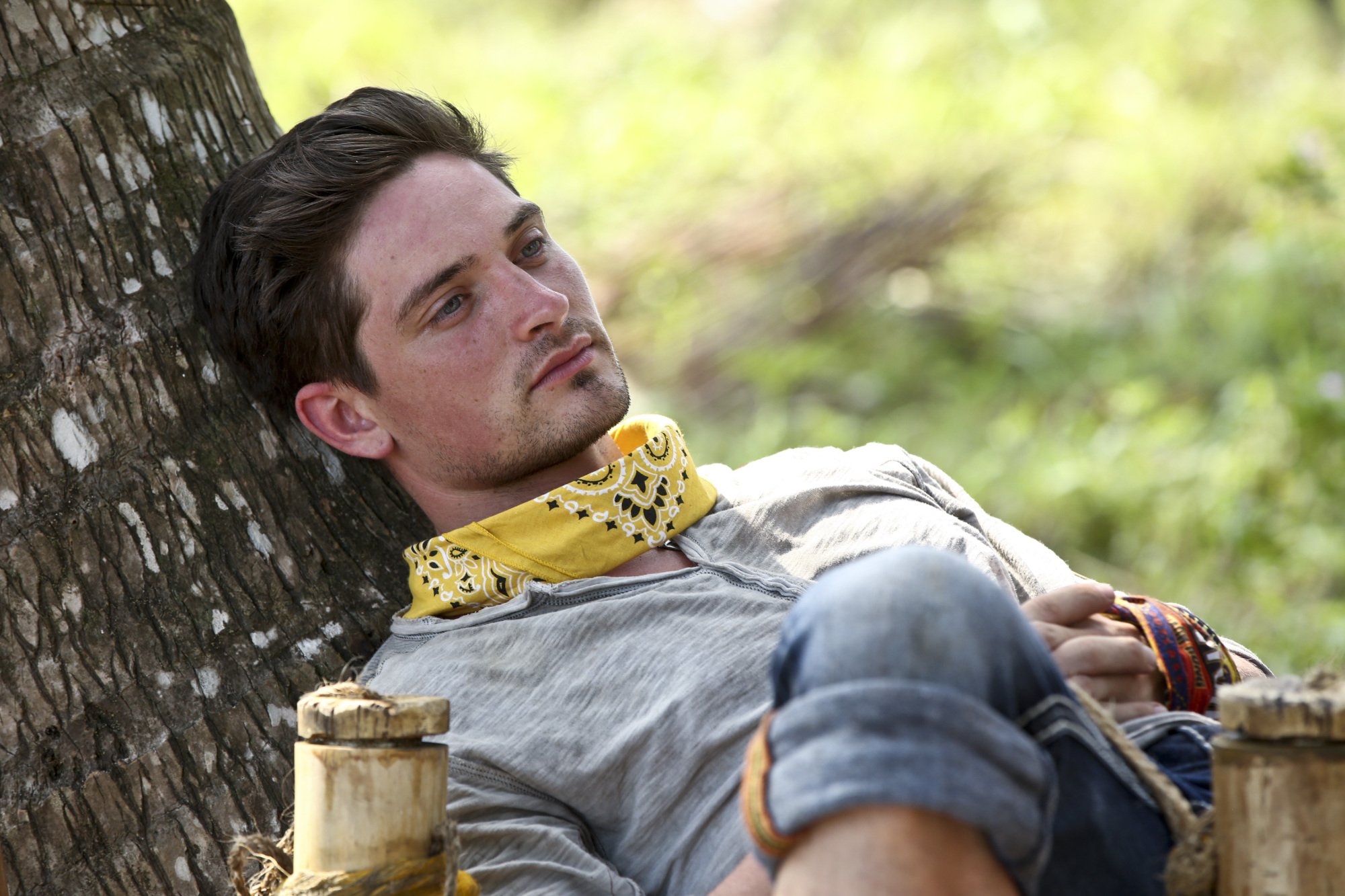 Caleb Reynolds, who starred in 'Survivor' Season 32 on CBS, wears a light gray henley shirt, rolled up jeans, and a yellow bandana around his neck.