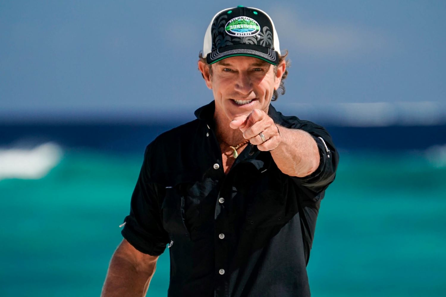 Jeff Probst, the host of 'Survivor 44' on CBS, wears a black button-up shirt with rolled-up sleeves and a black, white, and green 'Survivor' baseball cap while pointing at the camera.