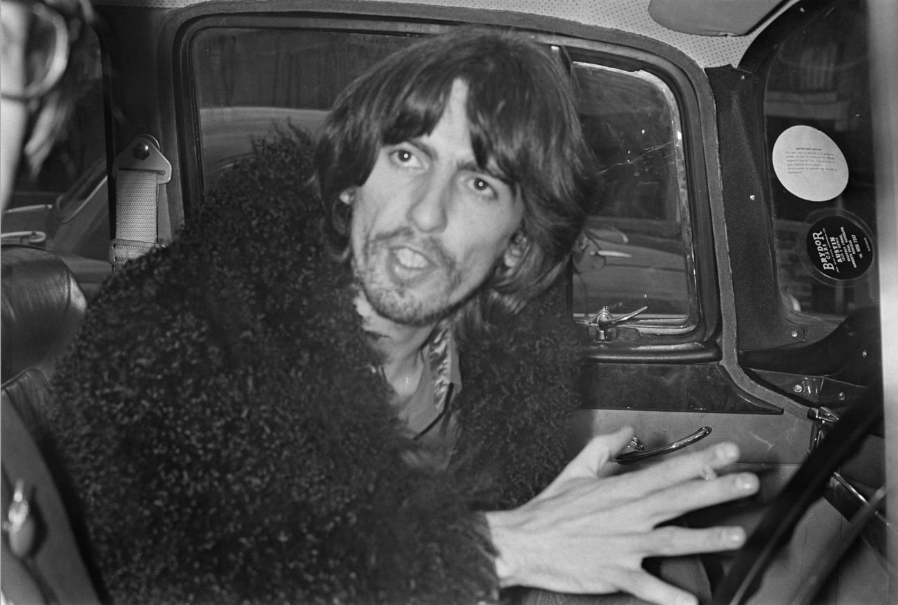 A black and white picture of George Harrison sitting in a car in a fuzzy coat.