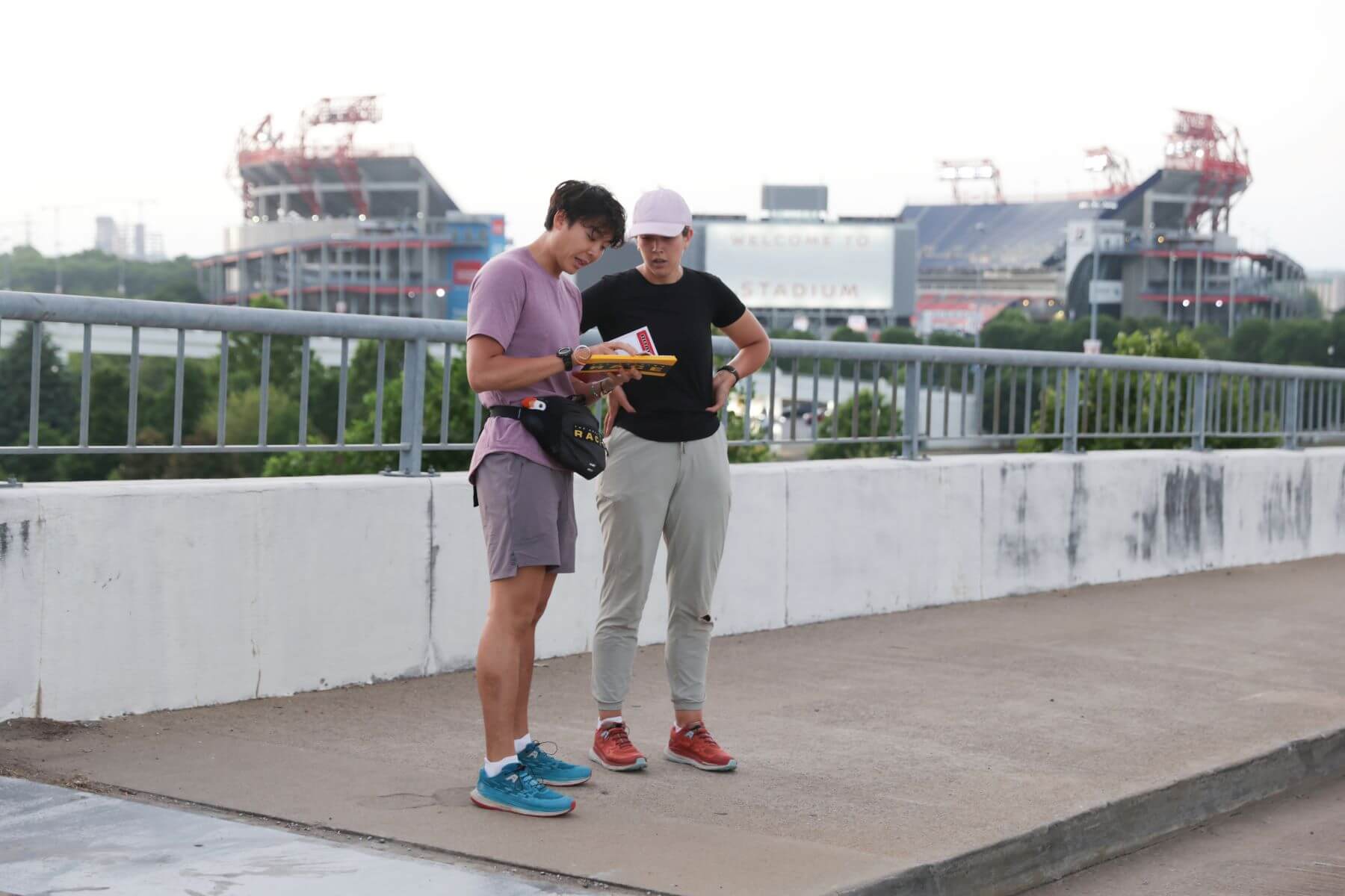 Derek Xiao and Claire Rehfuss read a clue while racing in the finale of 'The Amazing Race' Season 34 on CBS. Derek wears a light purple shirt, gray shorts, blue shoes, and a black fanny pack. Claire wears a black shirt, light gray pants, red shoes, and a light pink baseball cap.