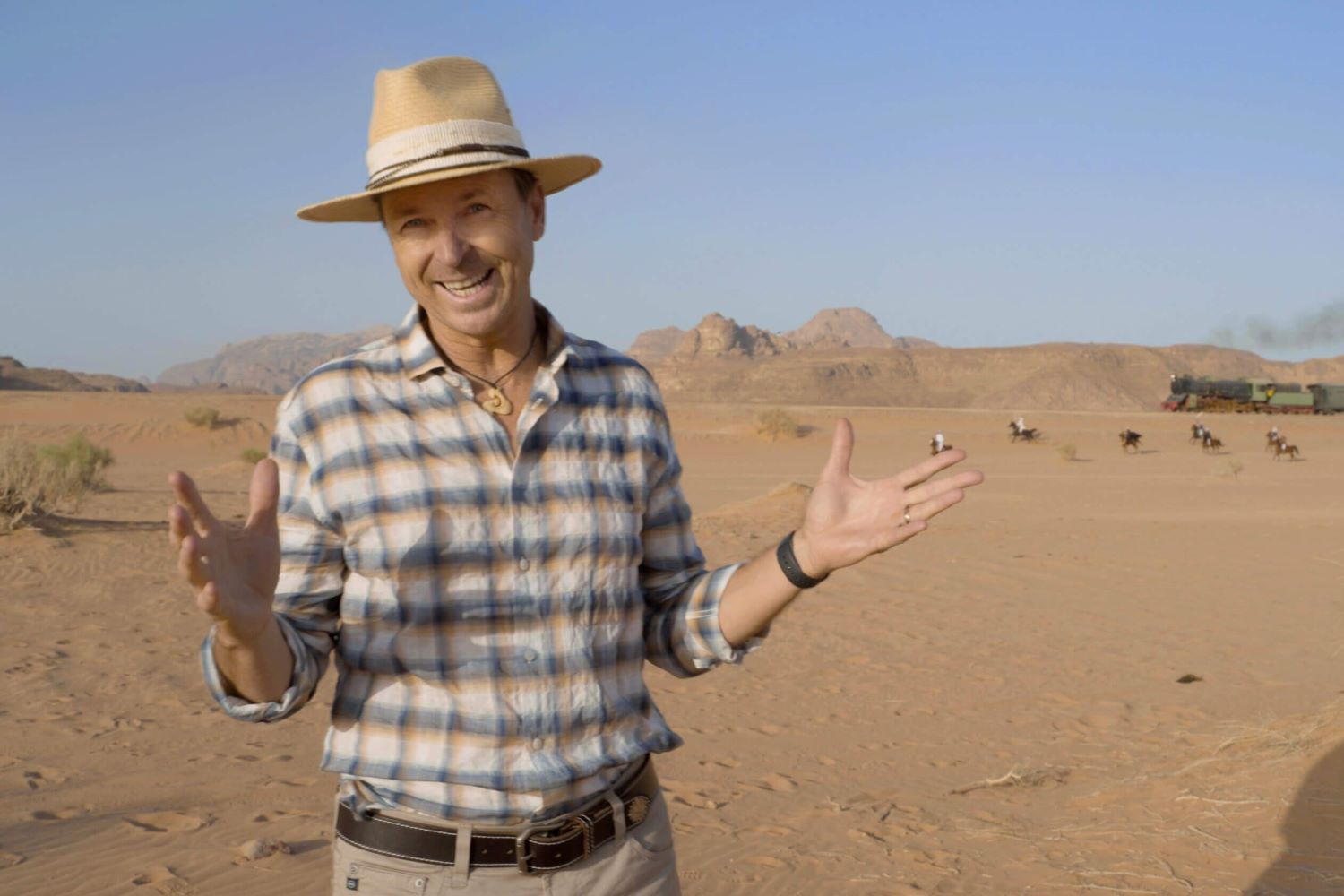 Phil Keoghan, the host of 'The Amazing Race' Season 35, appears in Jordan in season 34 wearing a blue, orange, and white plaid button-up shirt, tan pants, a brown belt, and a tan hat.