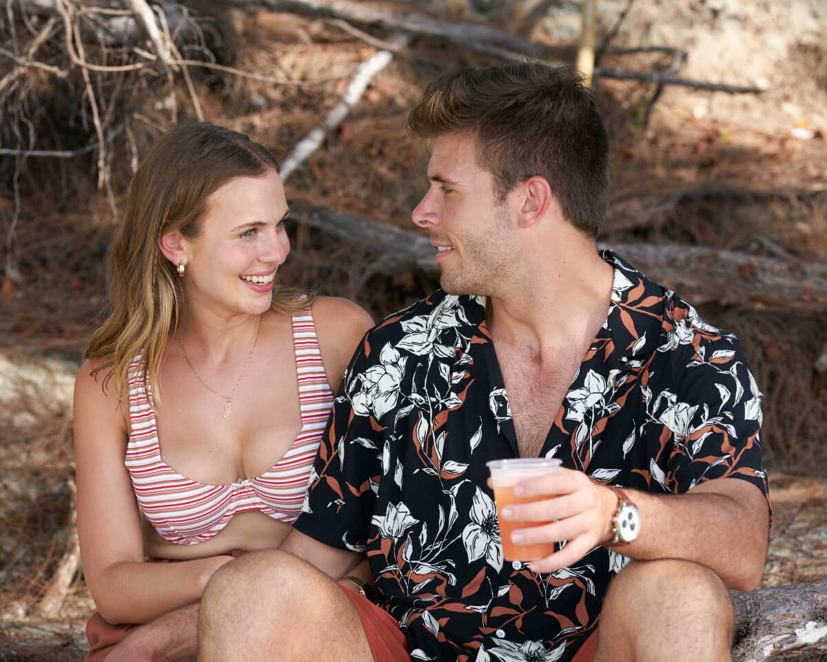 During The Bachelor Week 4, Jess Girod and Zach Shallcross talk on the beach. Jess wears a striped bikini top and Zach wears a floral shirt. 