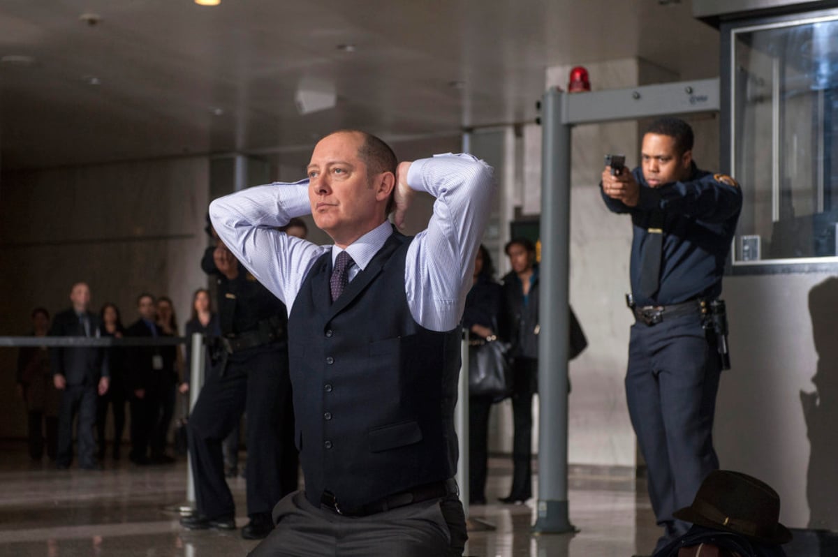 In The Blacklist pilot, Raymond Reddington kneels with his hands behind his head while a man points a gun at him.