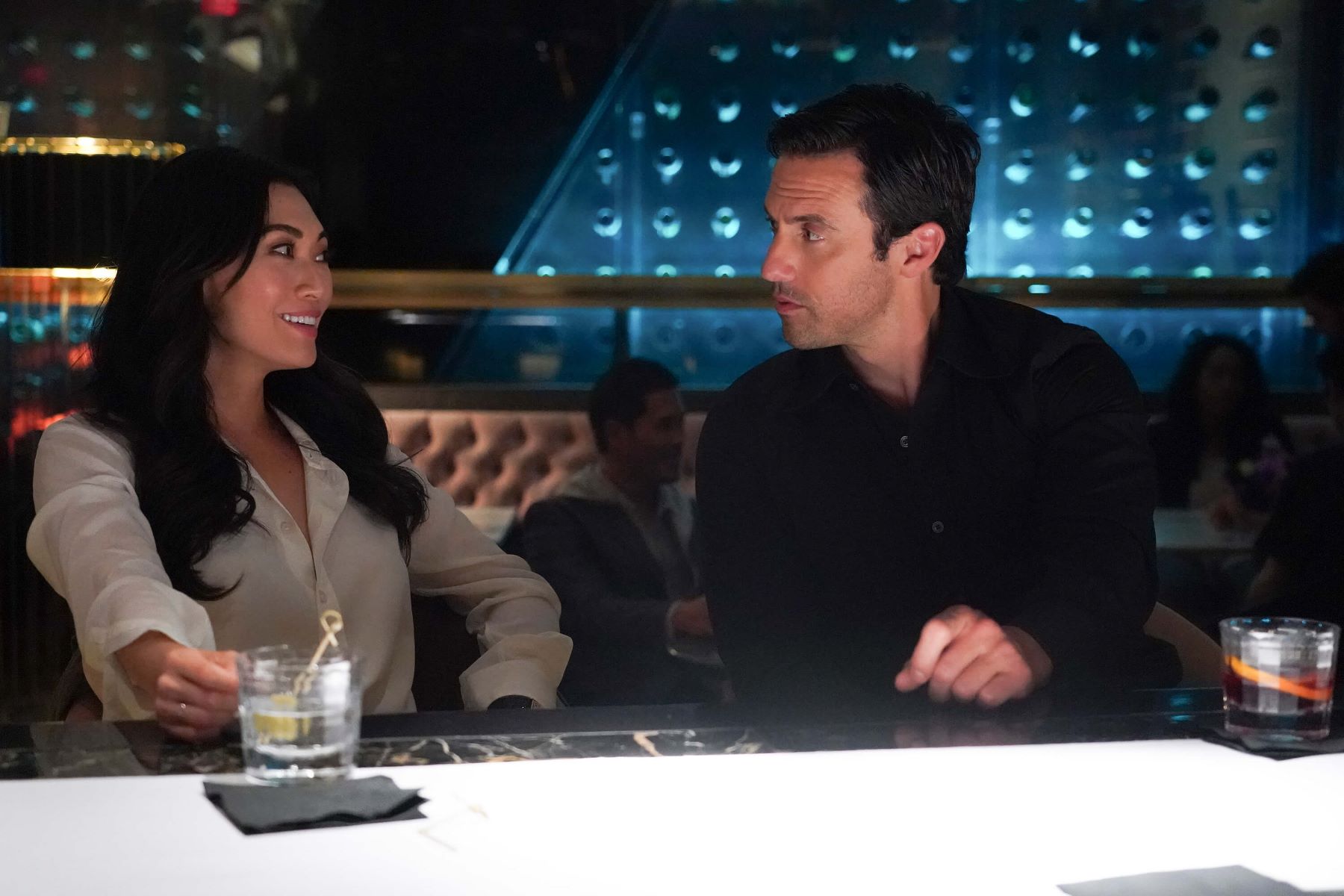 Catherine Haena Kim and Milo Ventimiglia, in character as Emma and Charlie in 'The Company You Keep' Season 1 Episode 1, share a scene at a bar. Emma wears a white long-sleeved button-up shirt. Charlie wears a black long-sleeved button-up shirt.