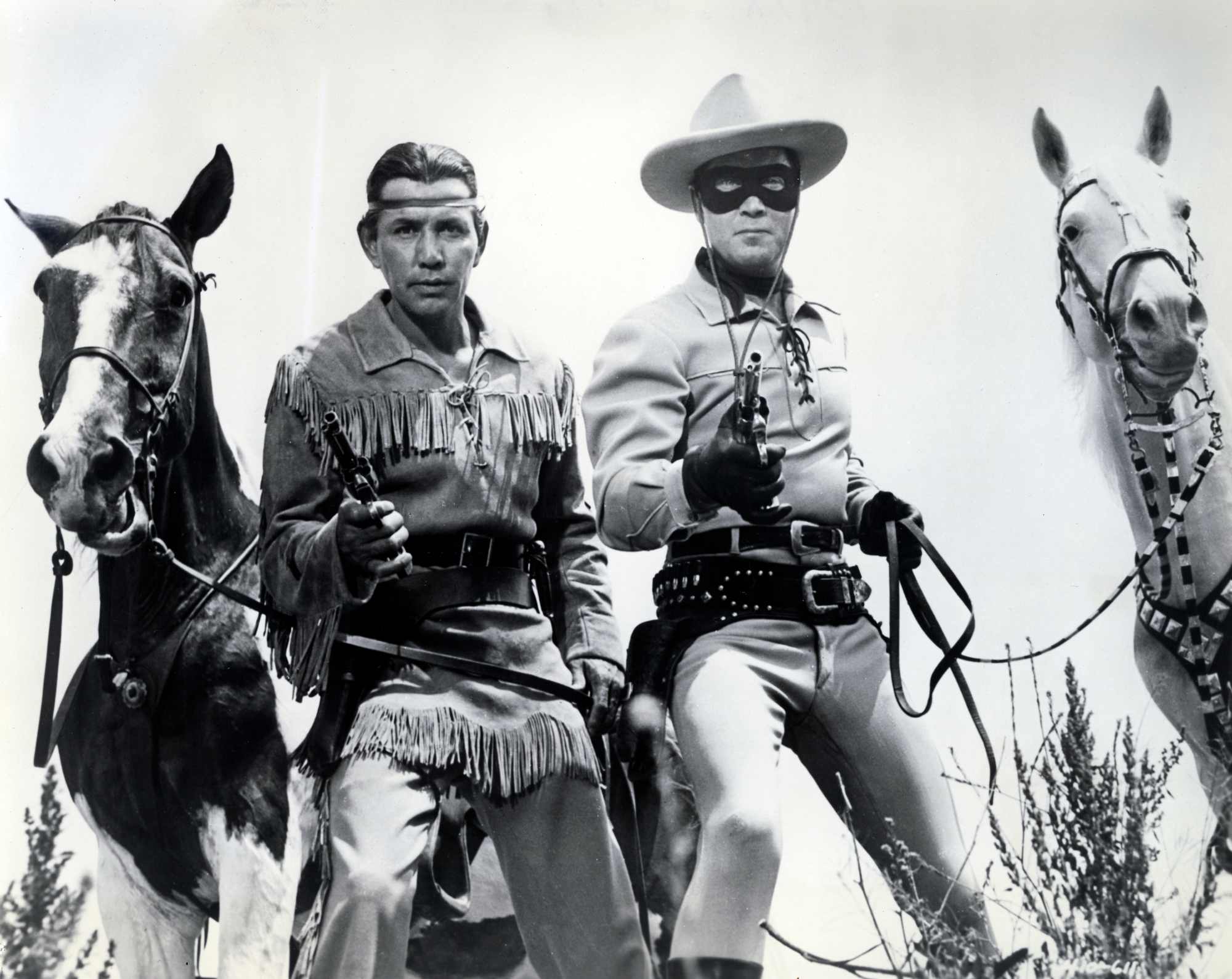 'The Lone Ranger' Jay Silverheels as Tonto and Clayton Moore as The Lone Ranger holding out their pistols, standing in front of their horses.