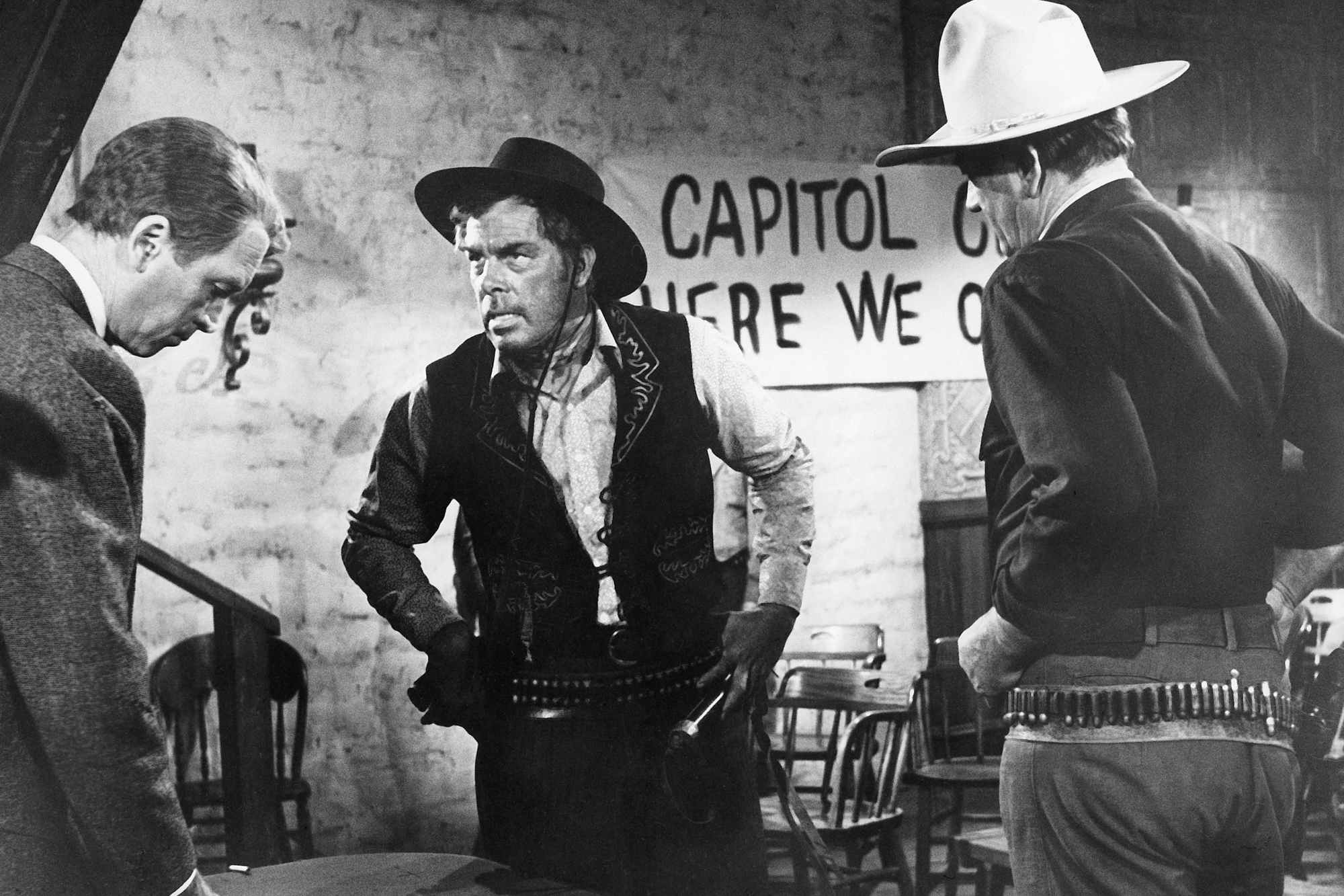 'The Man Who Shot Liberty Valance' James Stewart as Ransom Stoddard, Lee Marvin as Liberty Valance, and John Wayne as Tom Doniphon standing in front of a sign amongst tables. They're all wearing Western costumes.