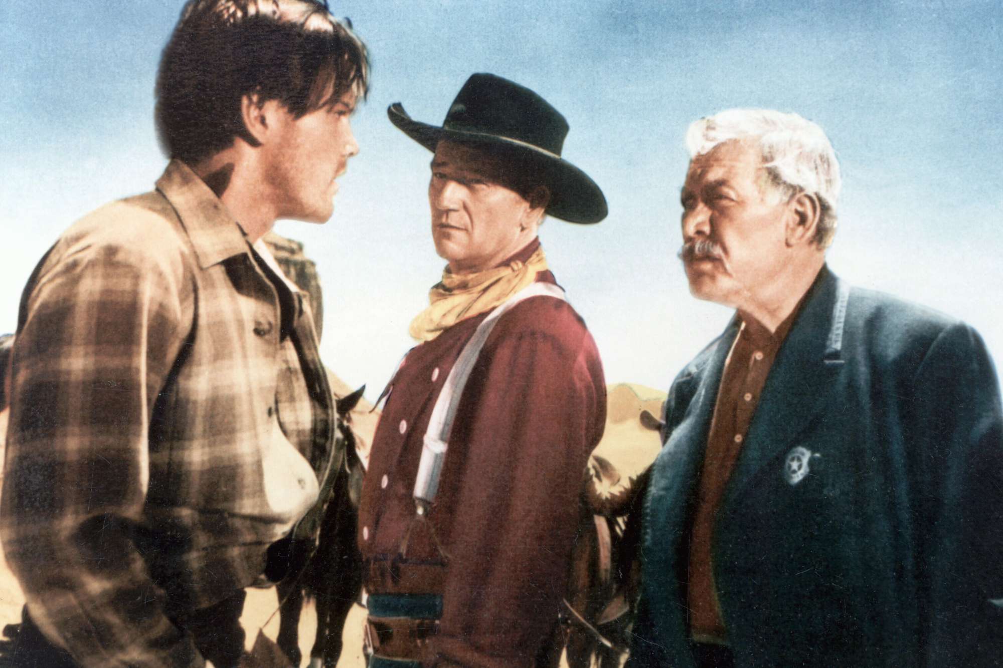 'The Searchers' Jeffrey Hunter as Martin Pawley, John Wayne as Ethan Edwards and Ward Bond as Rev. Capt. Samuel Johnston Clayton wearing Western costumes standing in the desert looking at one another