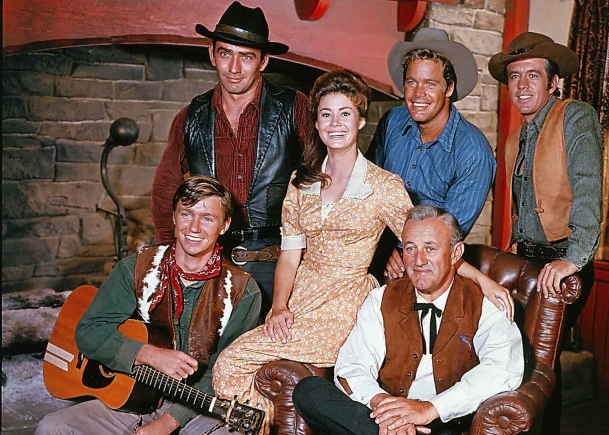 'The Virginians' Randy Boone as Randy Benton, James Drury as The Virginian, Roberta Shore as Betsy Garth, Doug McClure as Trampas, Clu Gulager as Emmett Ryker, Lee J. Cobb as Judge Henry Garth smiling around a sofa chair