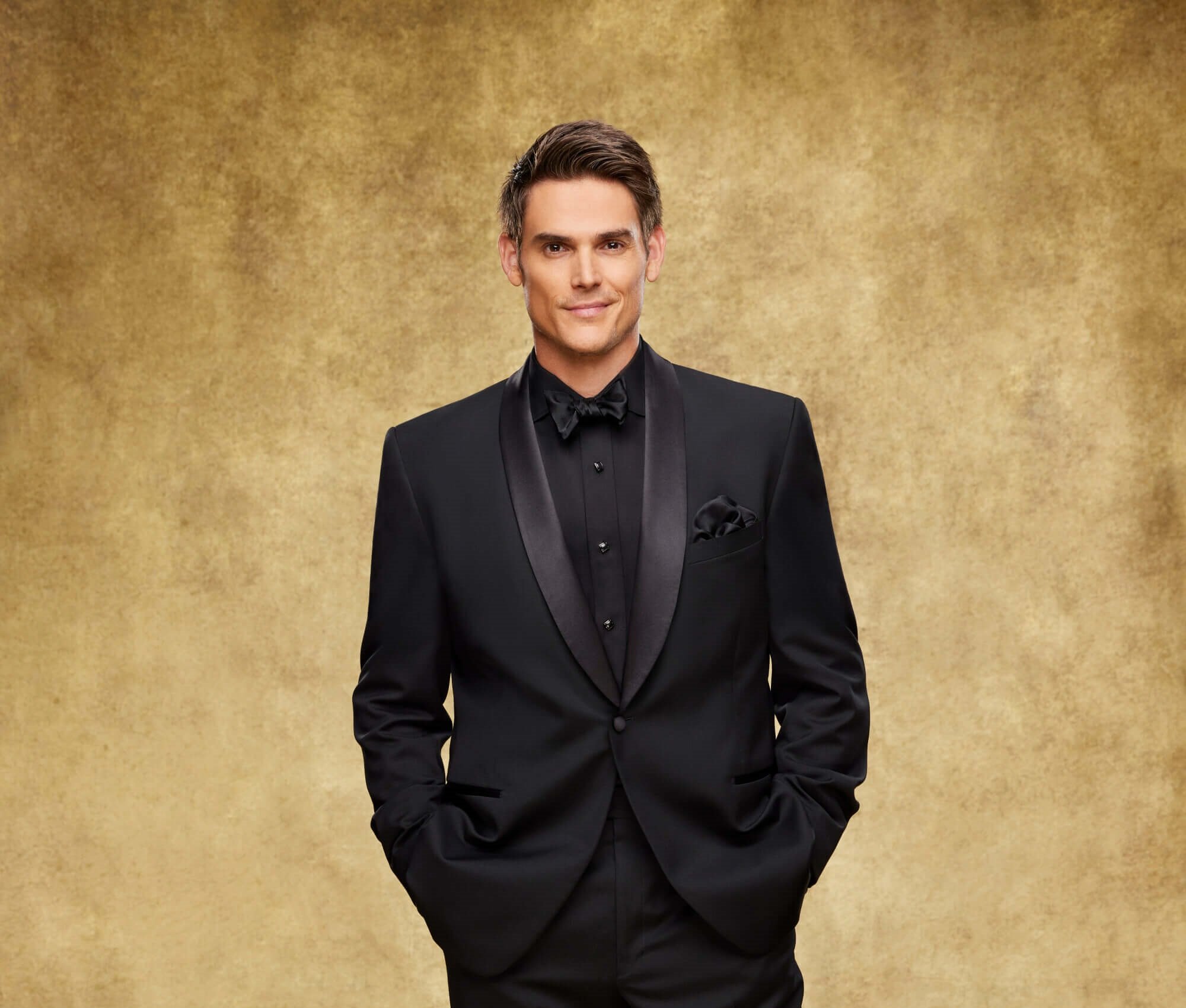 'The Young and the Restless' star Mark Grossman dressed in a black suit; posing in front of a gold backdrop.