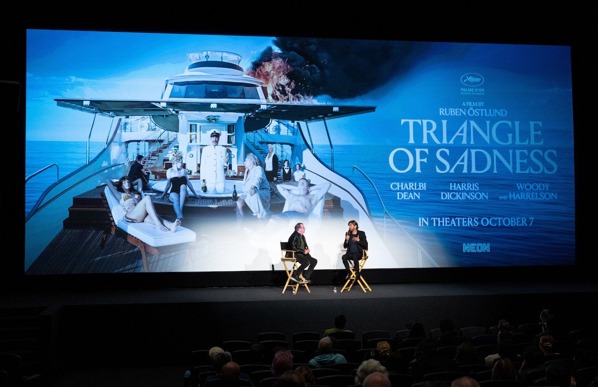 Film critic Pete Hammond and director Ruben Östlund sit in chairs in front of a screen that displays the "Triangle of Sadness" key art.