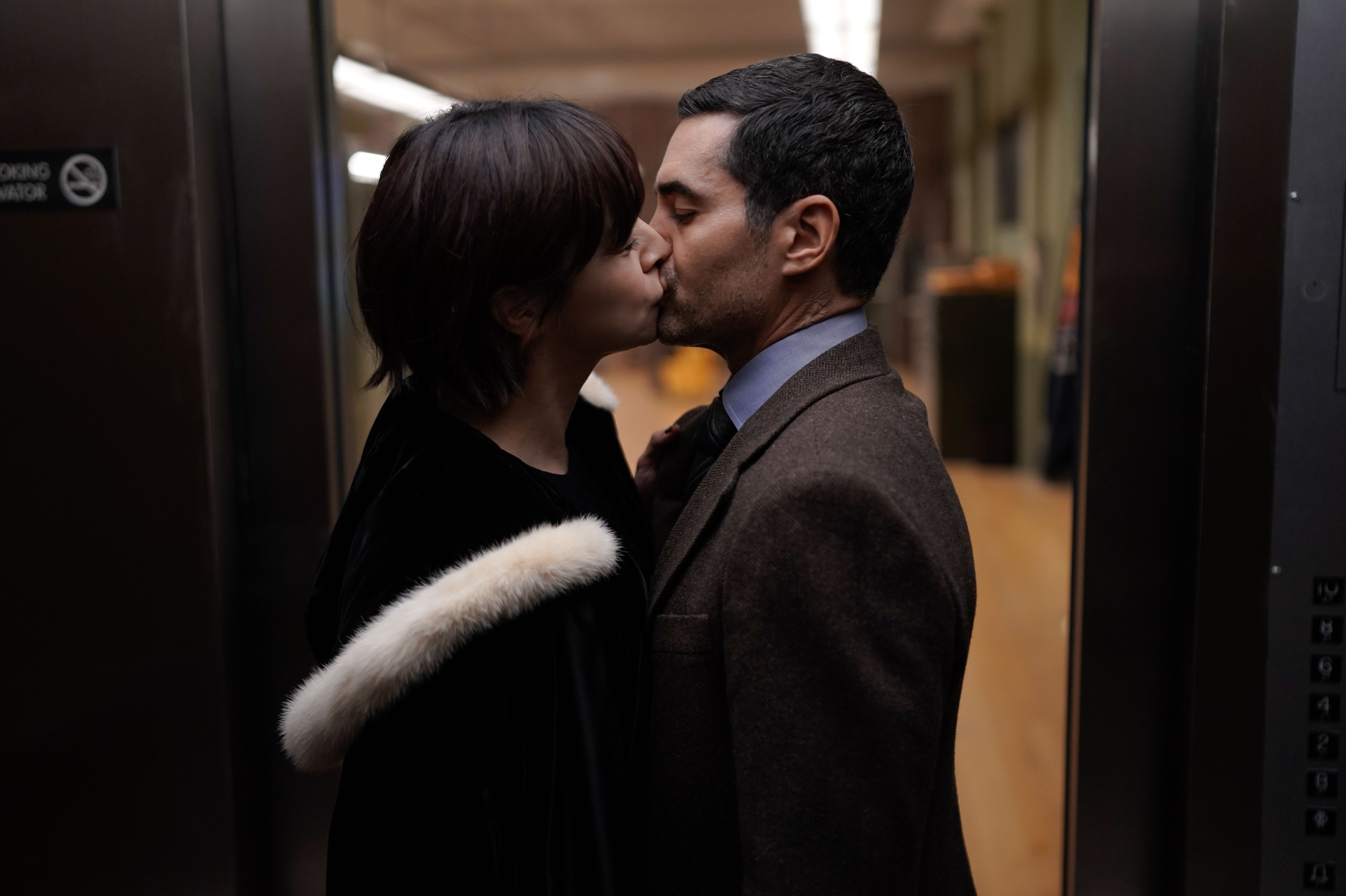 Julia Chan as Ava Green and Ramón Rodríguez as Will Trent kiss in 'Will Trent' Season 1 Episode 5. Ava wears a black coat with a white fur-lined hood. Will wears a dark brown three-piece suit.