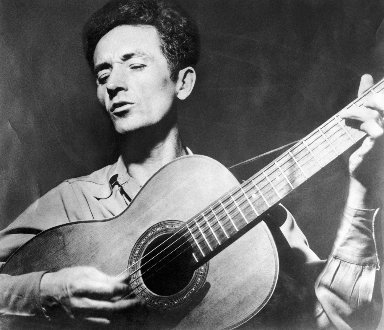 A black and white picture of Woody Guthrie playing guitar.