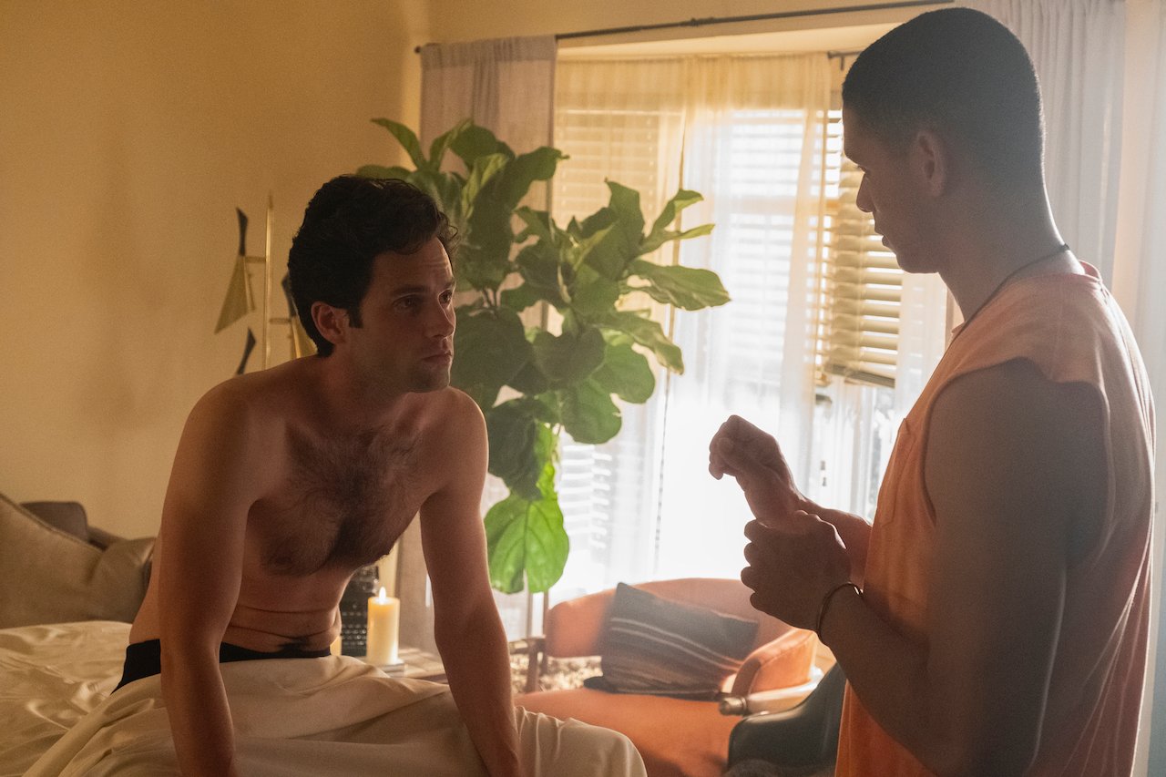Penn Badgley sits up shirtless in bed looking at Charlie Barnett in a scene for 'You'.