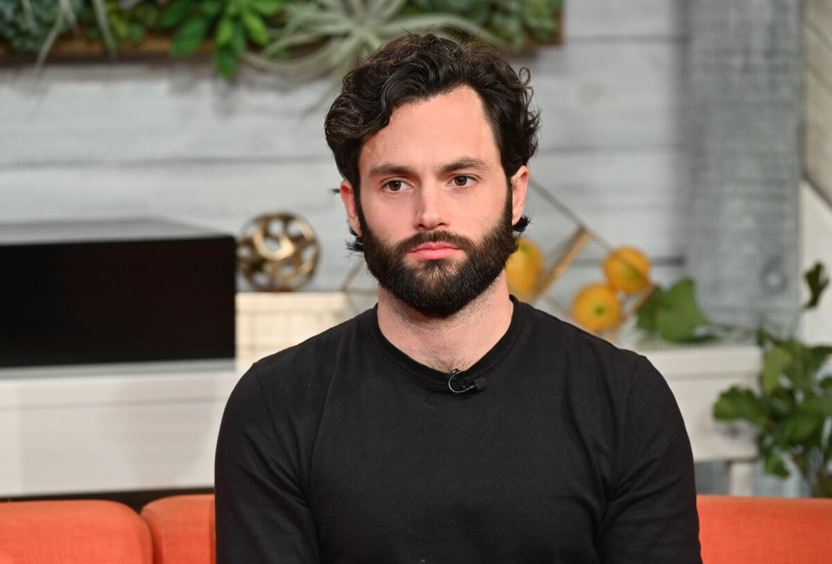 You star Penn Badgley sits on a couch wearing a black long-sleeved shirt.