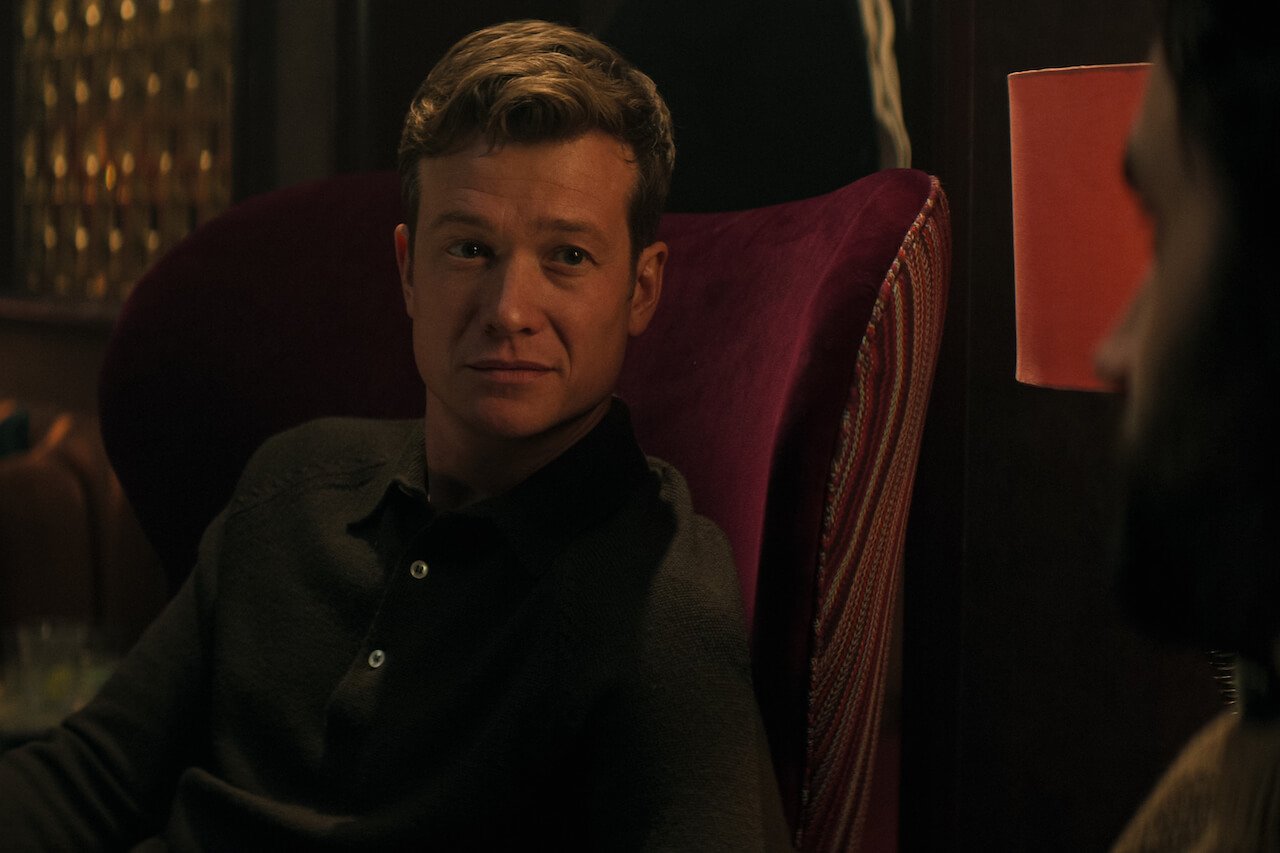 Ed Speleers as Rhys in 'You' Season 4 sits in a red chair.
