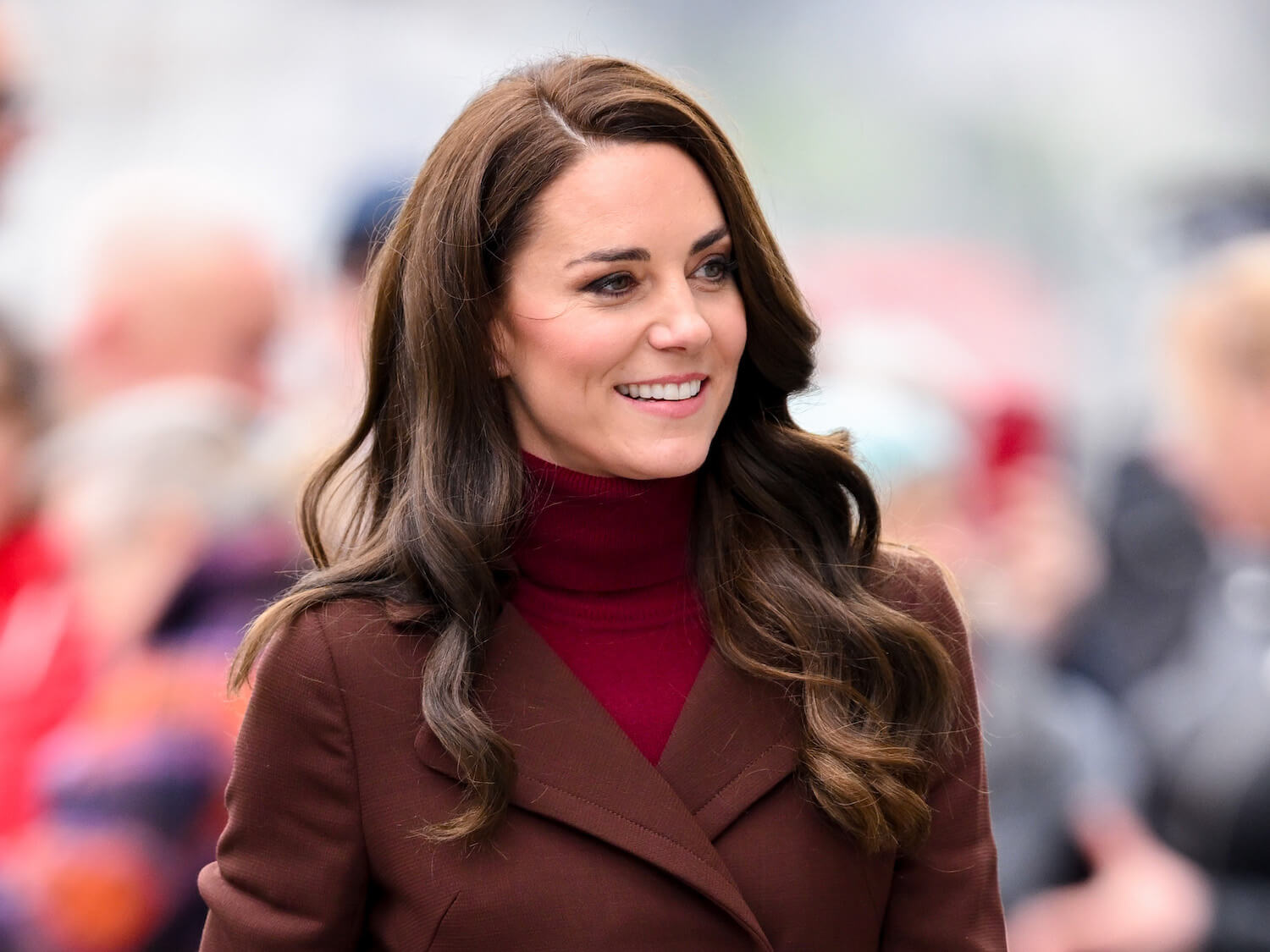 Kate Middleton, mom to three children, smiling in a brown coat and red sweater.
