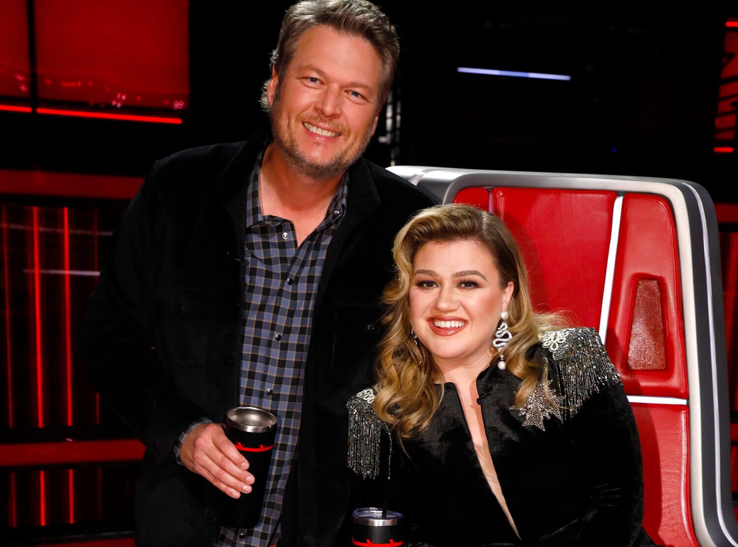 Blake Shelton stands beside Kelly Clarkson sitting in her chair on The Voice