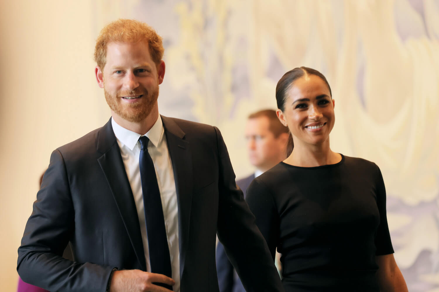 Prince Harry wears a suit and tie and smiles walking with Meghan Markle dressed in black