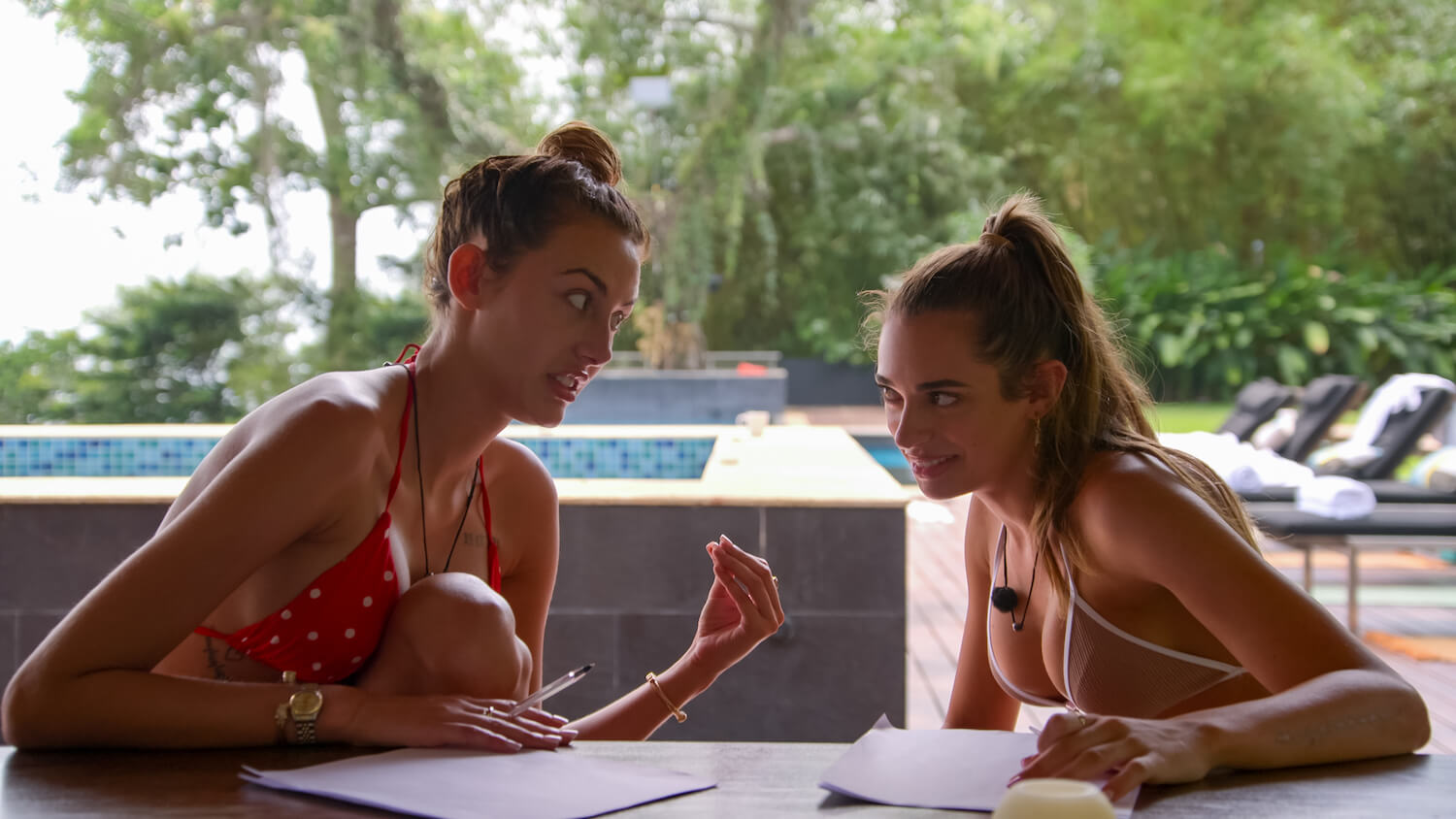 'Perfect Match' cast members Chloe Veitch and Georgia Hassarati sit at a table wearing bikinis in a production still from the series.