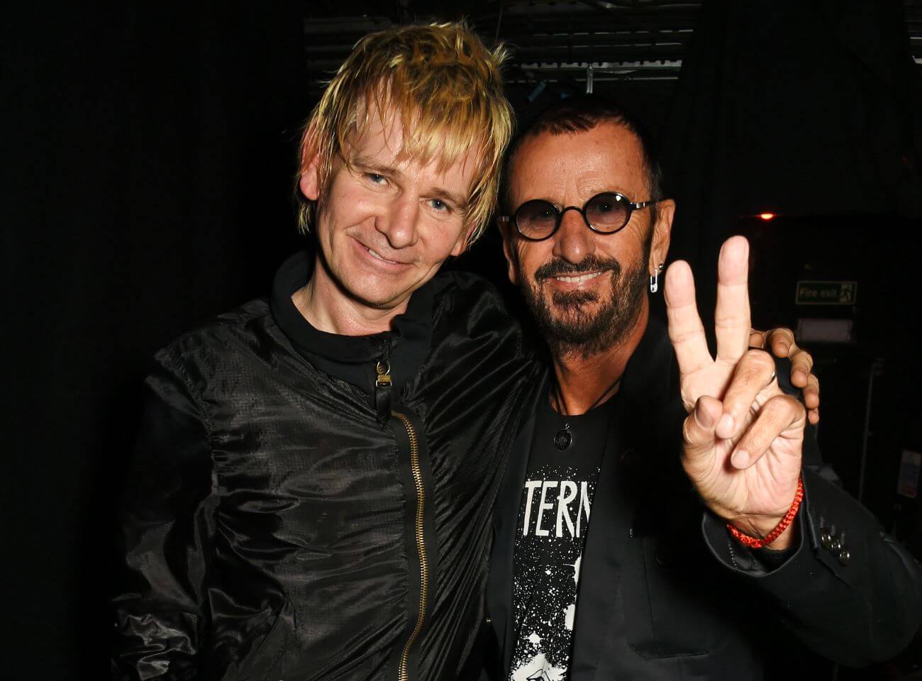Zak Starkey stands with his arm around Ringo Starr's shoulders. Ringo Starr holds up a peace sign.
