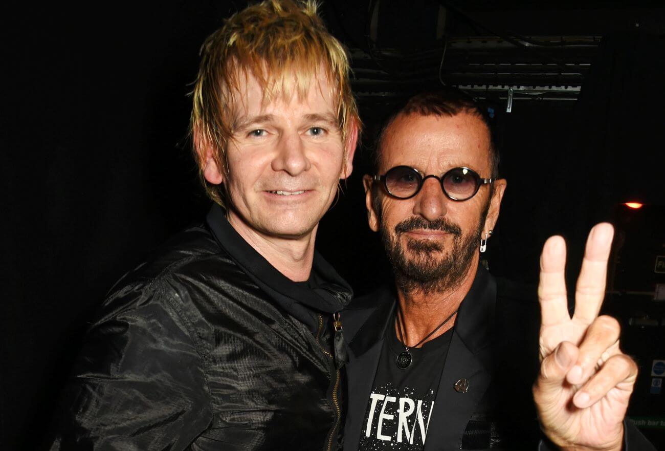 Zak Starkey stands with Ringo Starr. Ringo Starr holds up a peace sign.