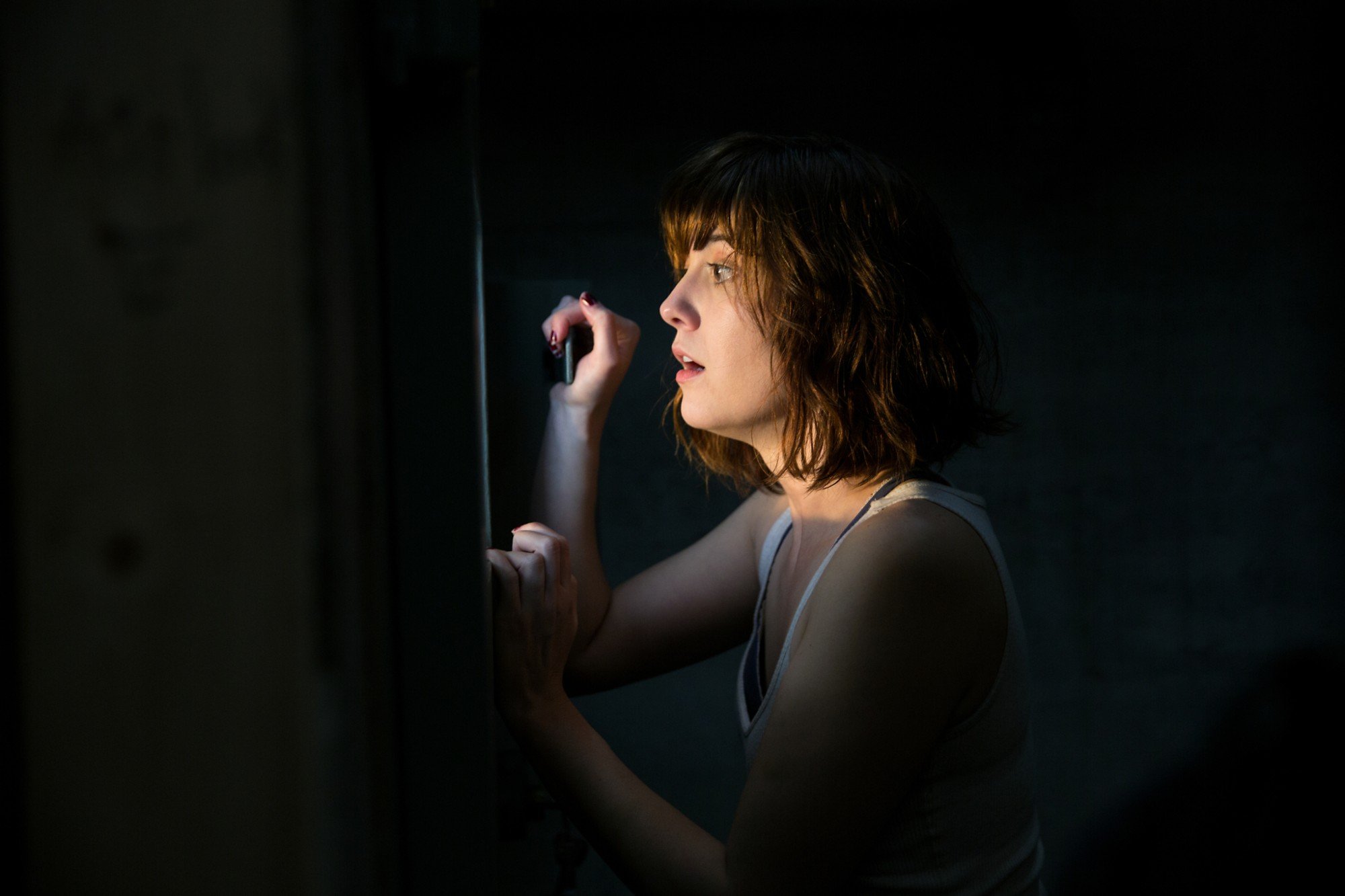 '10 Cloverfield Lane' Mary Elizabeth Winstead as Michelle standing in the dark, looking outside of a window, with a terrified look on her face.