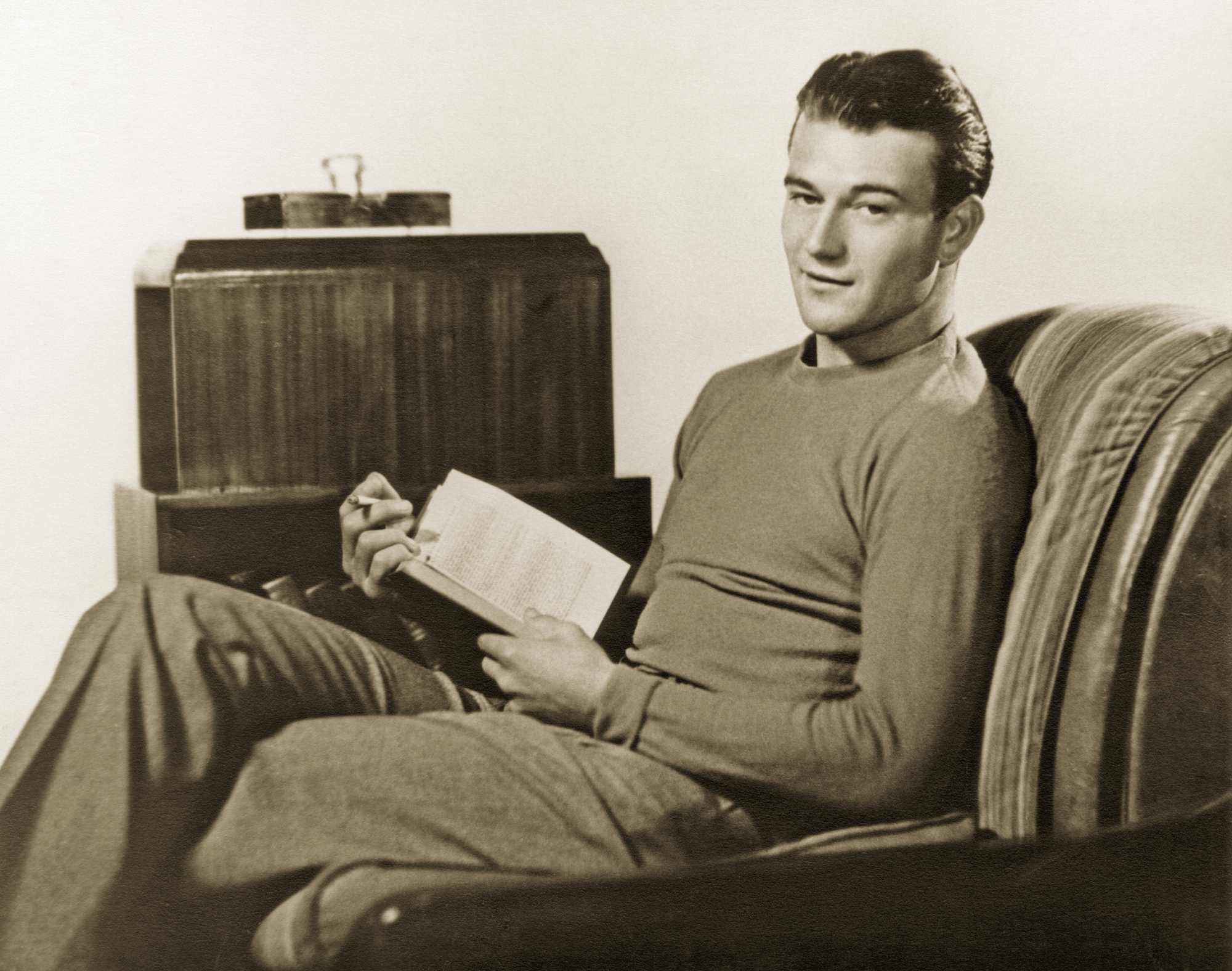 Actor John Wayne in a sepia tone picture, sitting in a sofa chair, holding a book.