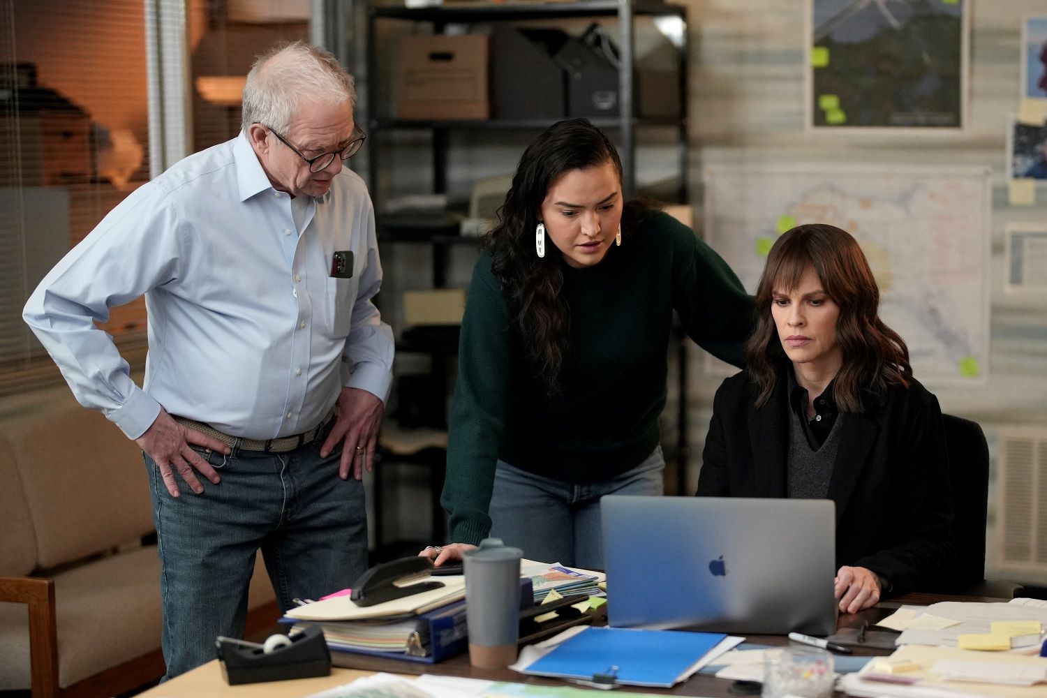 Jeff Perry as Stanley, Grace Dove as Roz, and Hilary Swank as Eileen share a scene in 'Alaska Daily' Episode 11. Stanley wears a light blue button-up shirt and jeans. Roz wear a dark green sweater and jeans. Eileen wears a black coat over a gray sweater vest over a black button-up shirt.