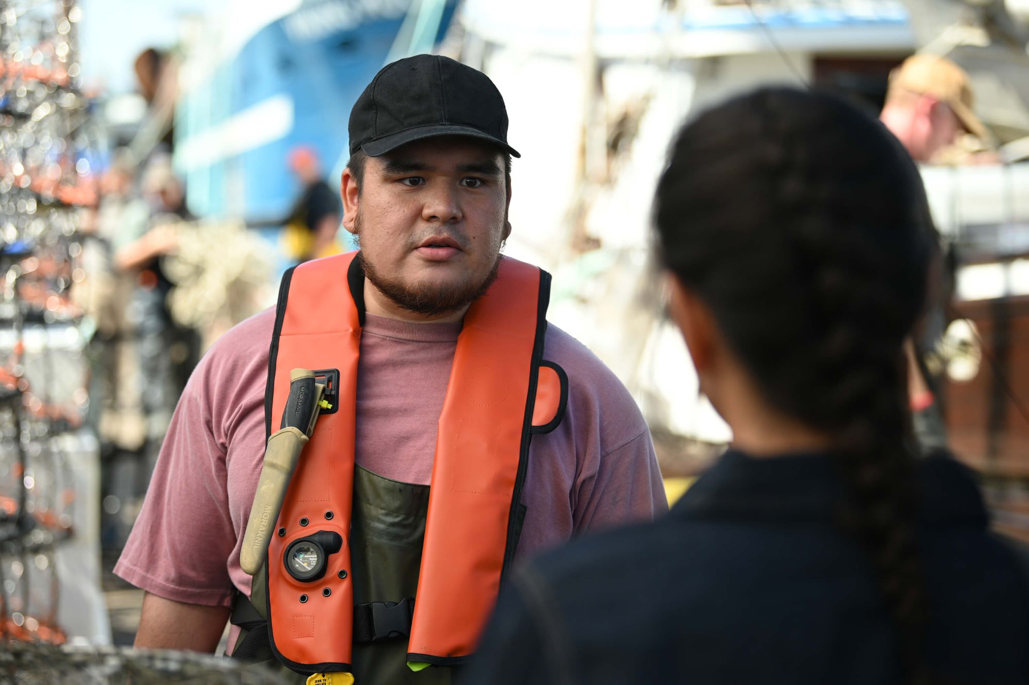 Kam Kozak, in character as Toby Crenshaw in 'Alaska Daily' Episode 2, wears an orange life vest over a pink shirt and a black baseball cap.