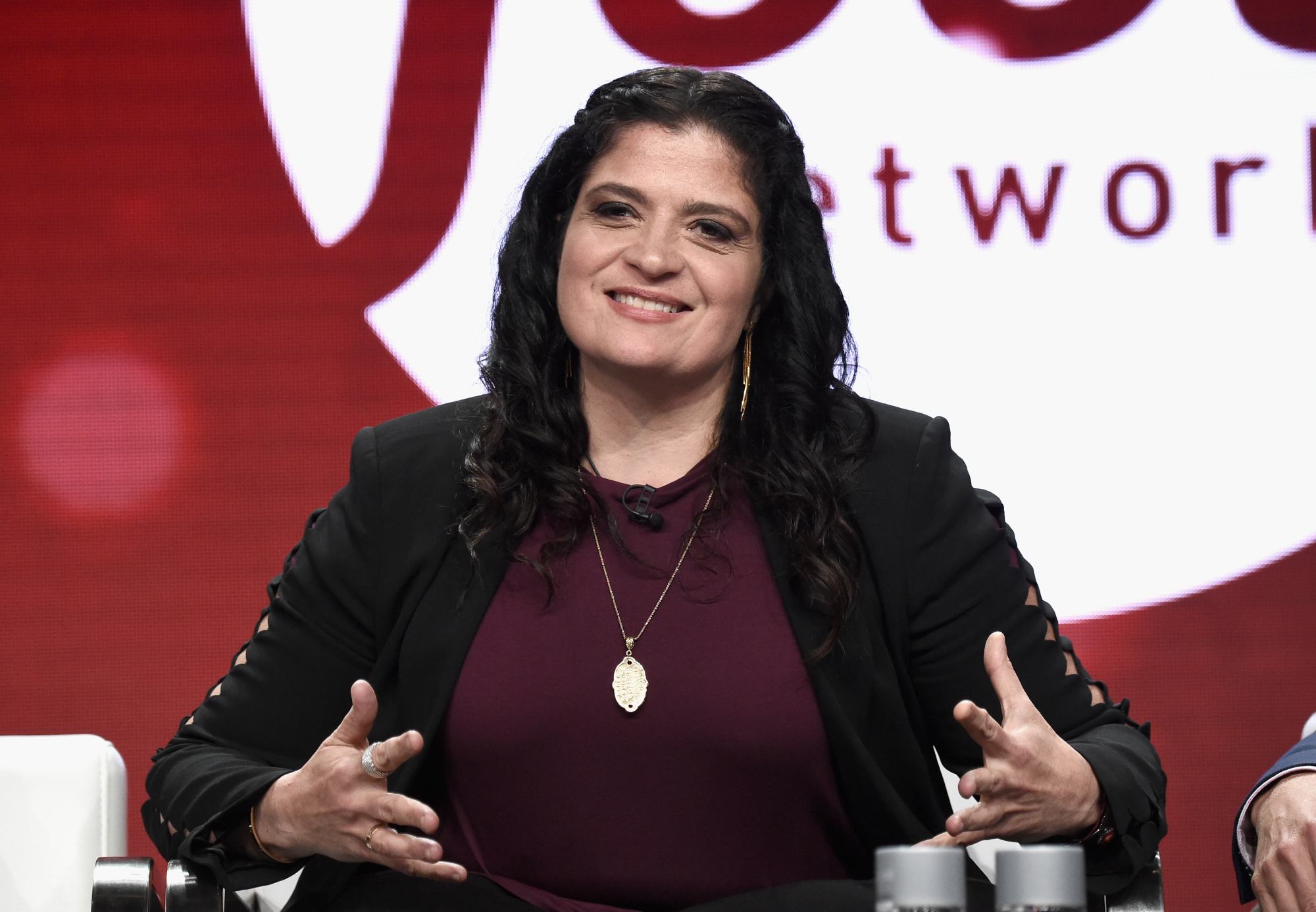 Alex Guarnaschelli smiling, talking with her hands in front of the Food Network logo.
