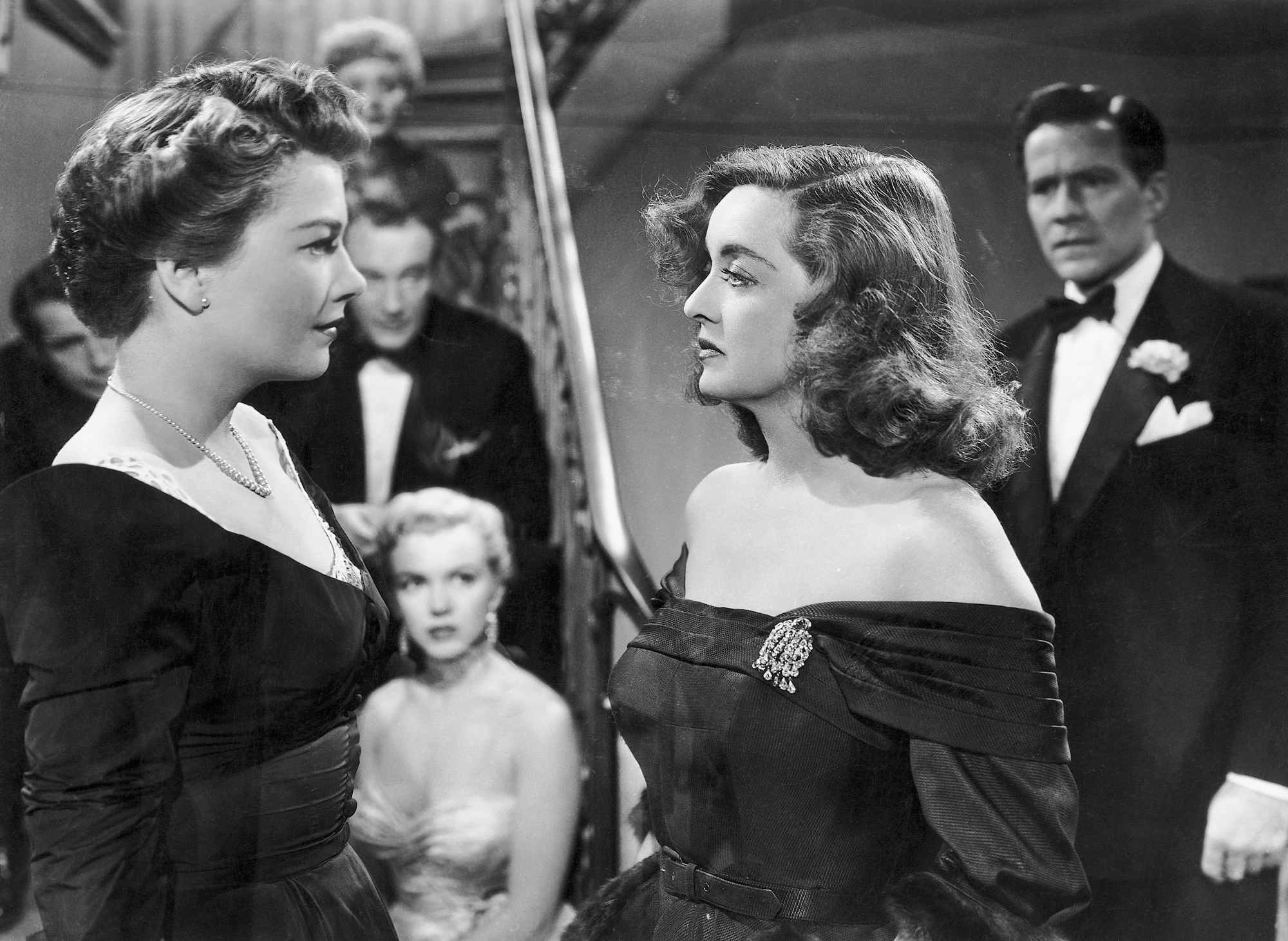 'All About Eve' Bette Davis as Margo and Anne Baxter as Eve looking at one another intensely with onlookers standing behind them on a staircase.