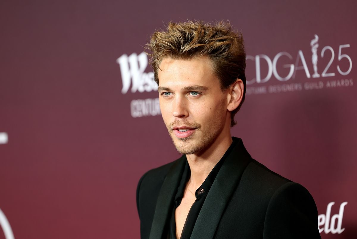 Austin Butler smiles for the camera in front of a maroon backdrop.
