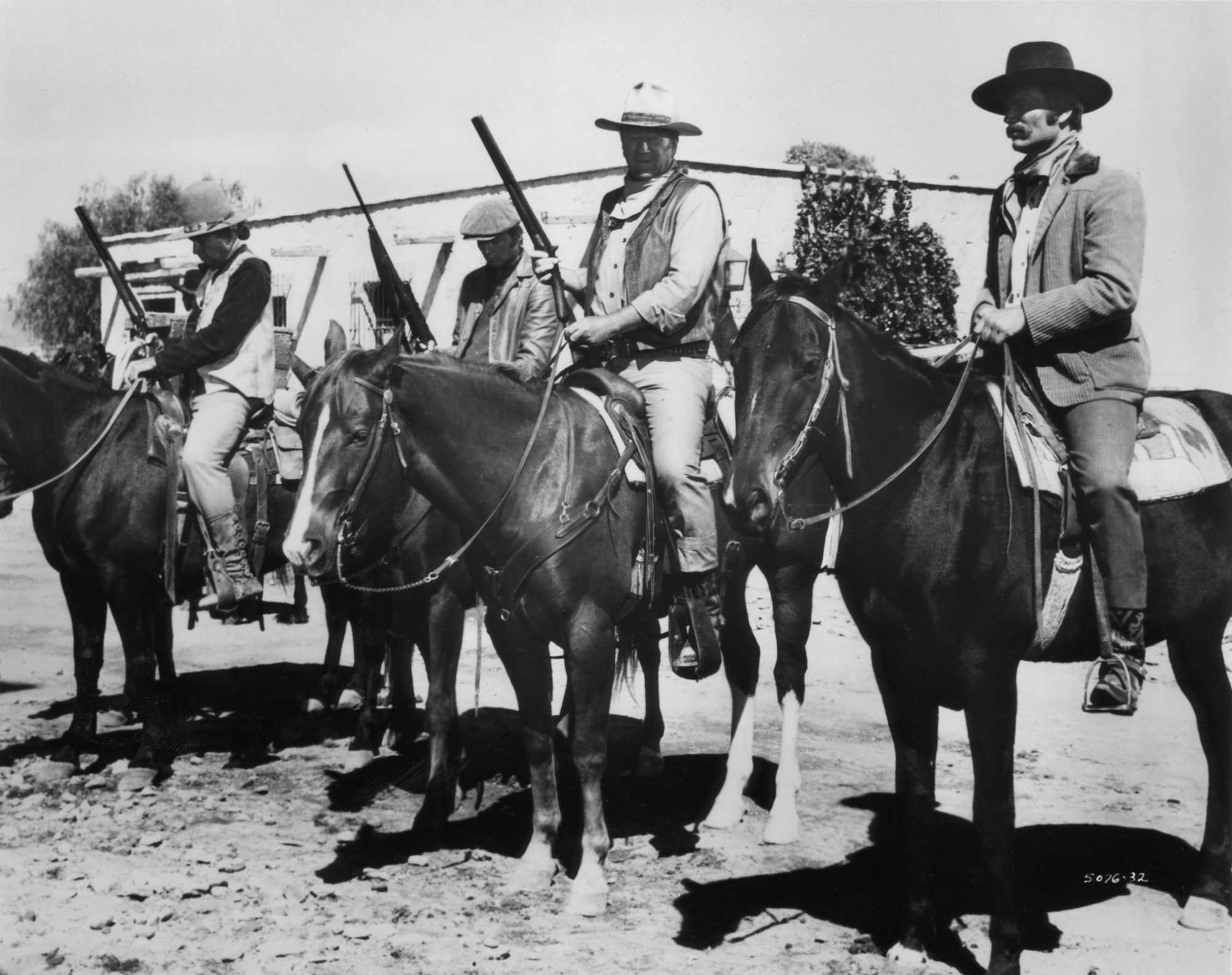 'Big Jake' Bruce Cabot as Sam Sharpnose, Christopher Mitchum as Michael McCandles, John Wayne as Jacob McCandles, and Patrick Wayne as James McCandles on their horses, holding guns in front of a shack.
