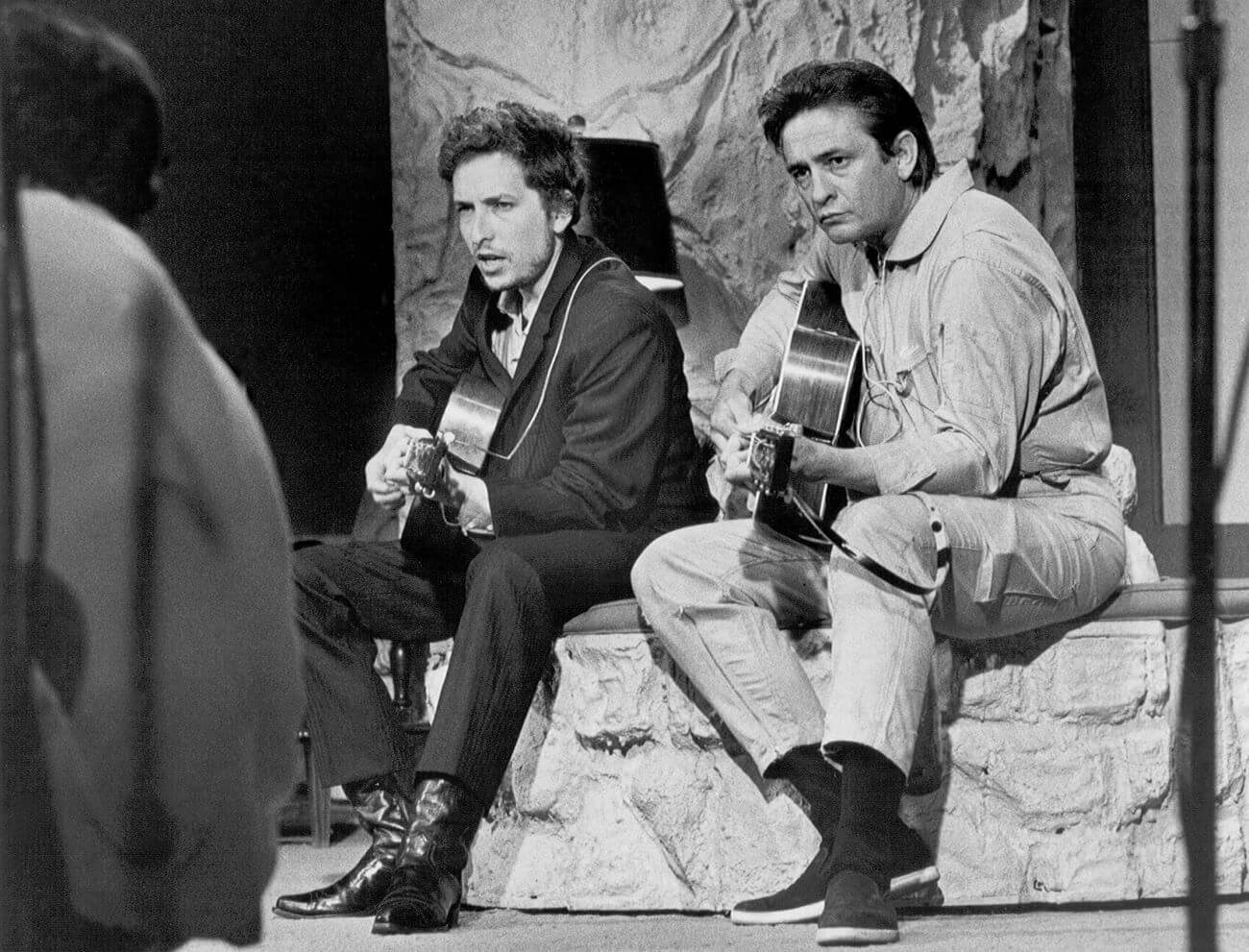 A black and white picture of Bob Dylan and Johnny Cash sitting on a stone ledge together with guitars.