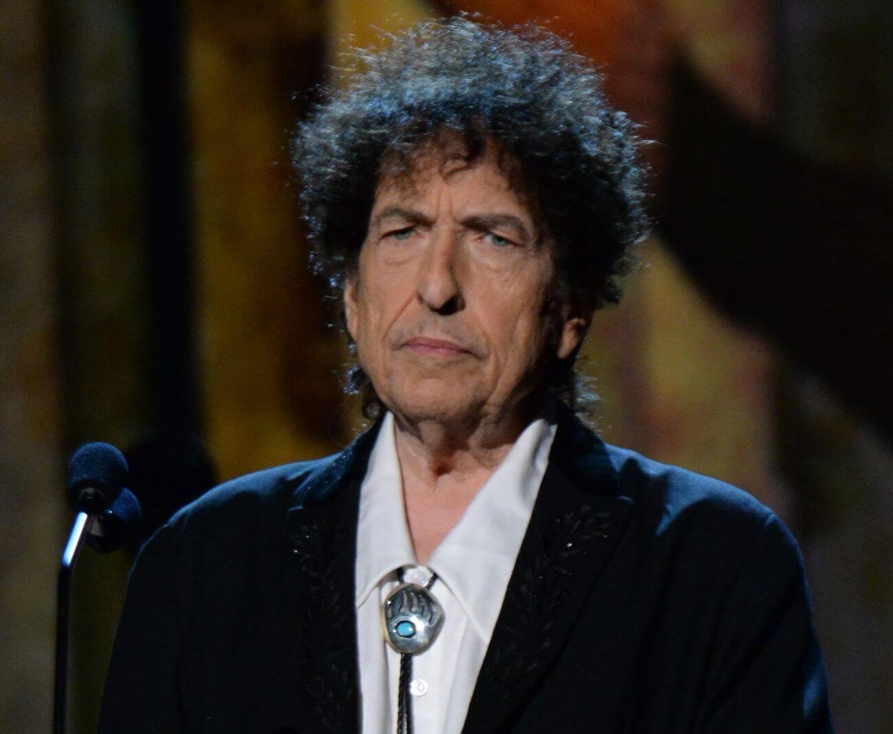 Bob Dylan wears a bolo tie and stands in front of a microphone.