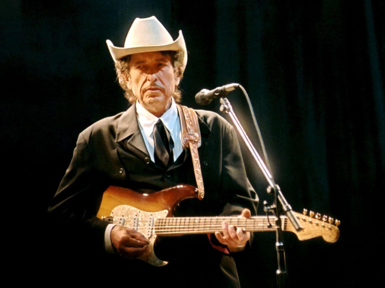 Bob Dylan wears a cowboy hat and holds a guitar while standing in front of a microphone.